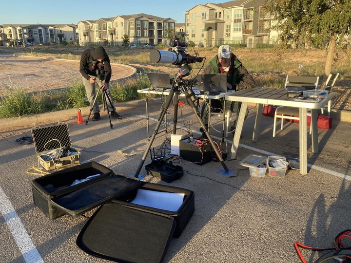 On a parking lot near white and grey three-floor residential buildings two men are hunched over telescopes and computers, respectively. Square-sided equipment bags lie open on the pavement. The shadows cast on the ground are long, indicating it is the beginning or end of a long day. 