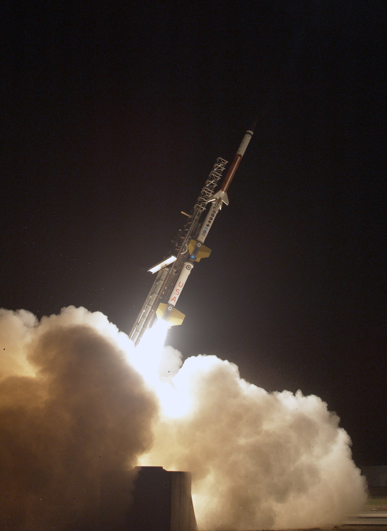 a 3 stage sounding rocket launches off a rail against a inky black sky. In the foreground plumes of white smoke are lit up by the rocket's ignition