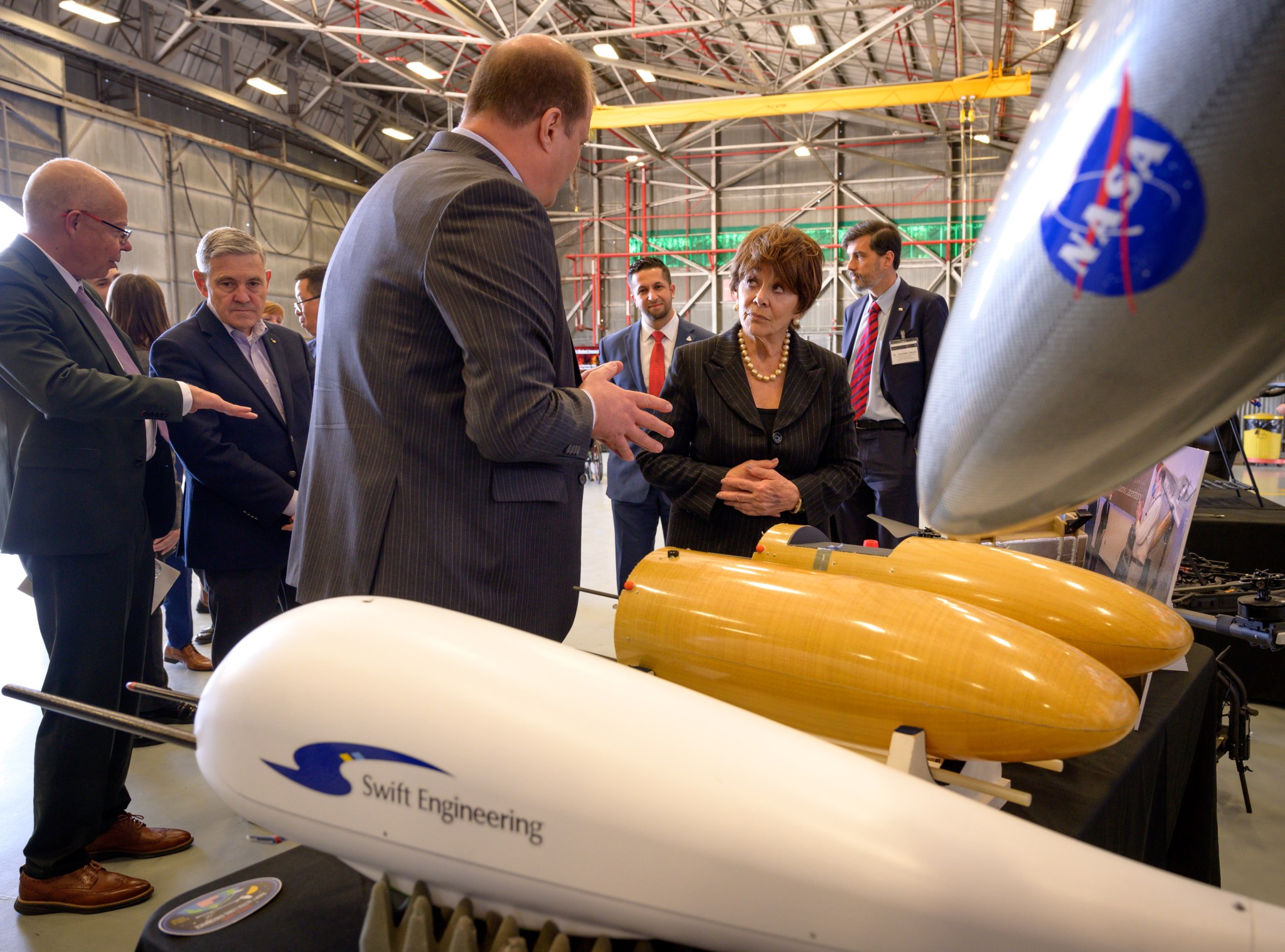 A man in a gray suit with back to camera talks with a woman in a dark suit next to a display of drone components: two orange conical pieces, a white component with a Swift Engineering logo, and a gray piece with NASA logo.