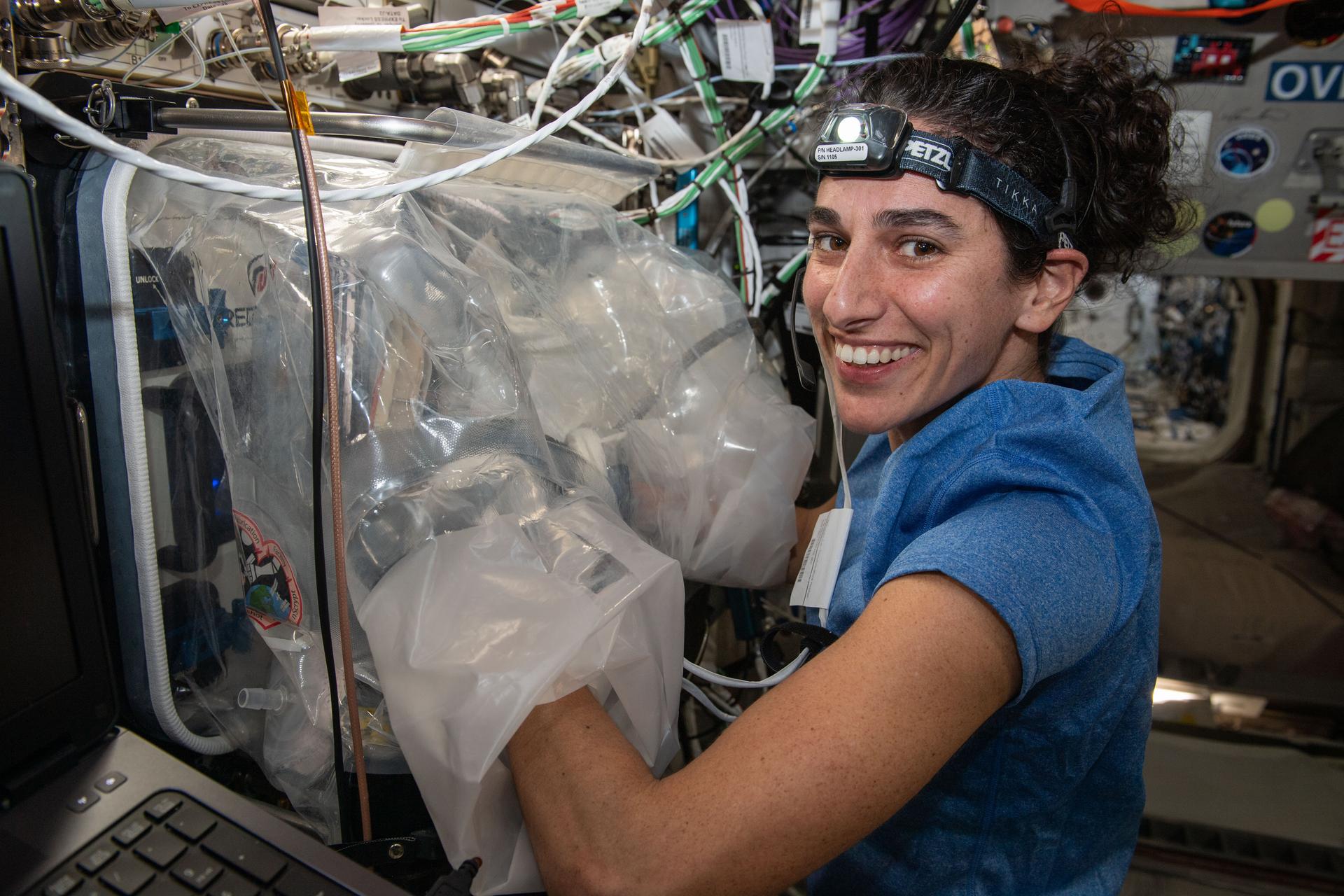 Moghbeli’s arms are inserted into large plastic gloves that are connected to a clear flexible plastic glovebag attached to the wall of the space station. Moghbeli is wearing a blue shirt and a headlamp. She is looking at the camera over her shoulder and smiling.