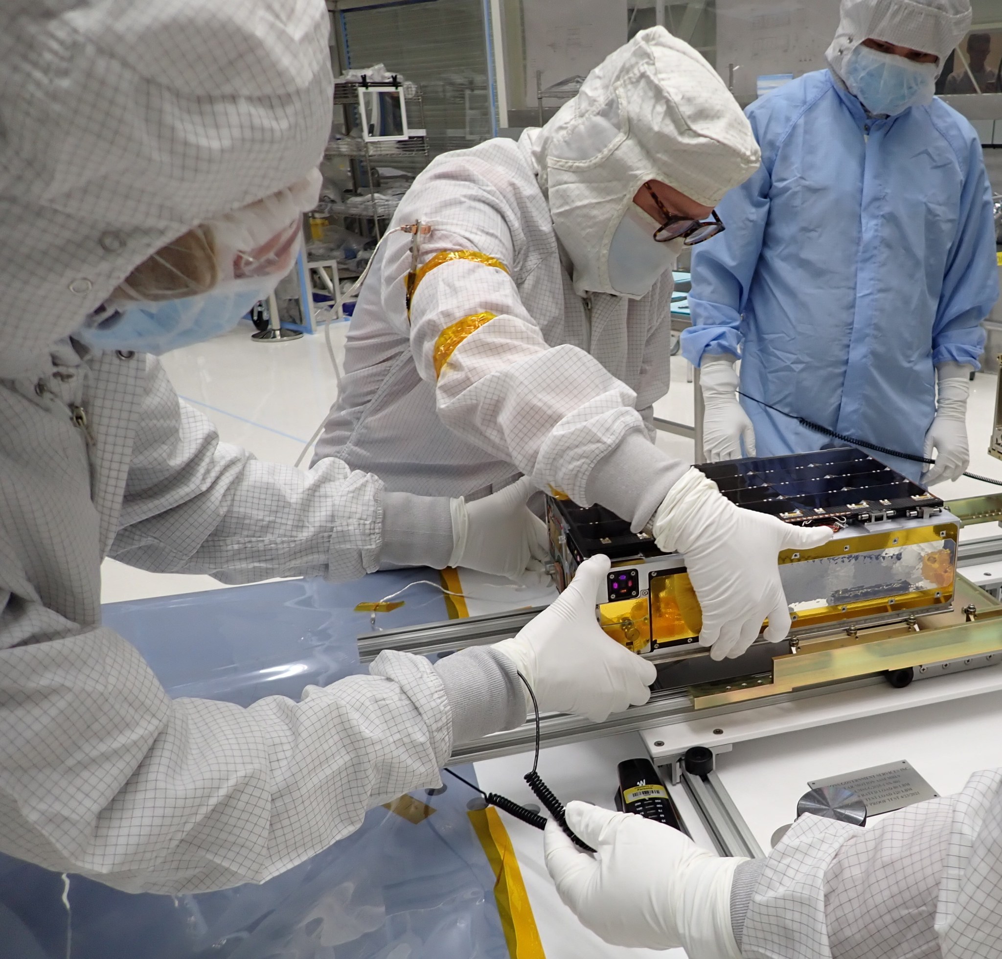 Several individuals dressed in protective clothing, masks and gloves attach a rectangular box to rails.