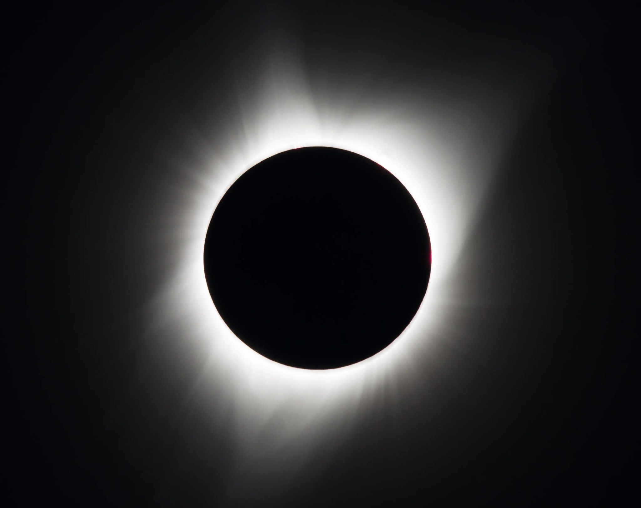 Against a black background is a total solar eclipse. In the middle is a black circle – the Moon. Surrounding it are white streams of wispy light, streaming out into the sky.