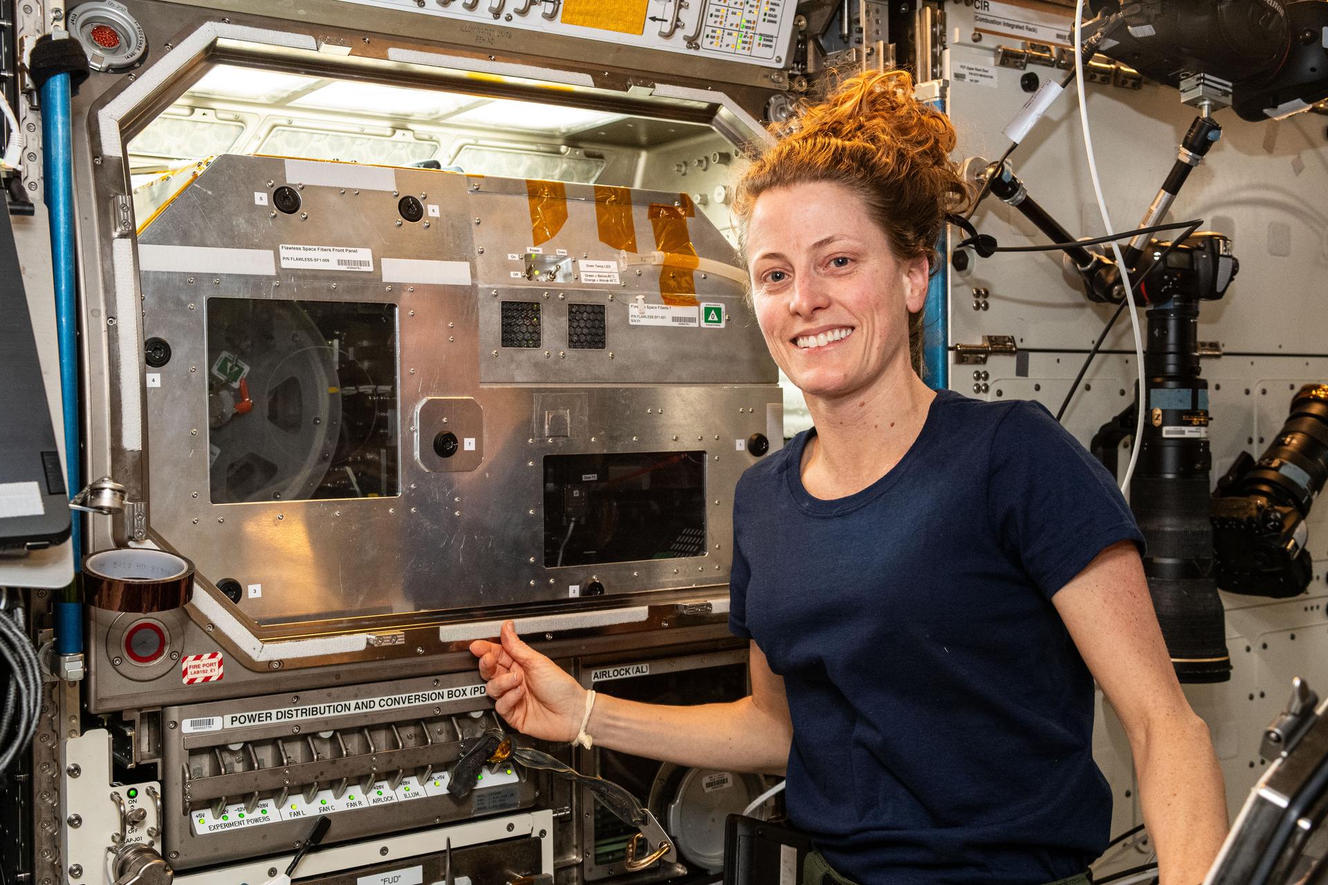 O’Hara, wearing a dark blue sleeveless t-shirt, smiles at the camera. Her right hand is touching the front of a large silver box built into a wall, with lights above it and a clear panel that reveals part of a circular sample holder that resembles an old film reel. There is a panel of switches below her hand and several large cameras on the wall behind her.