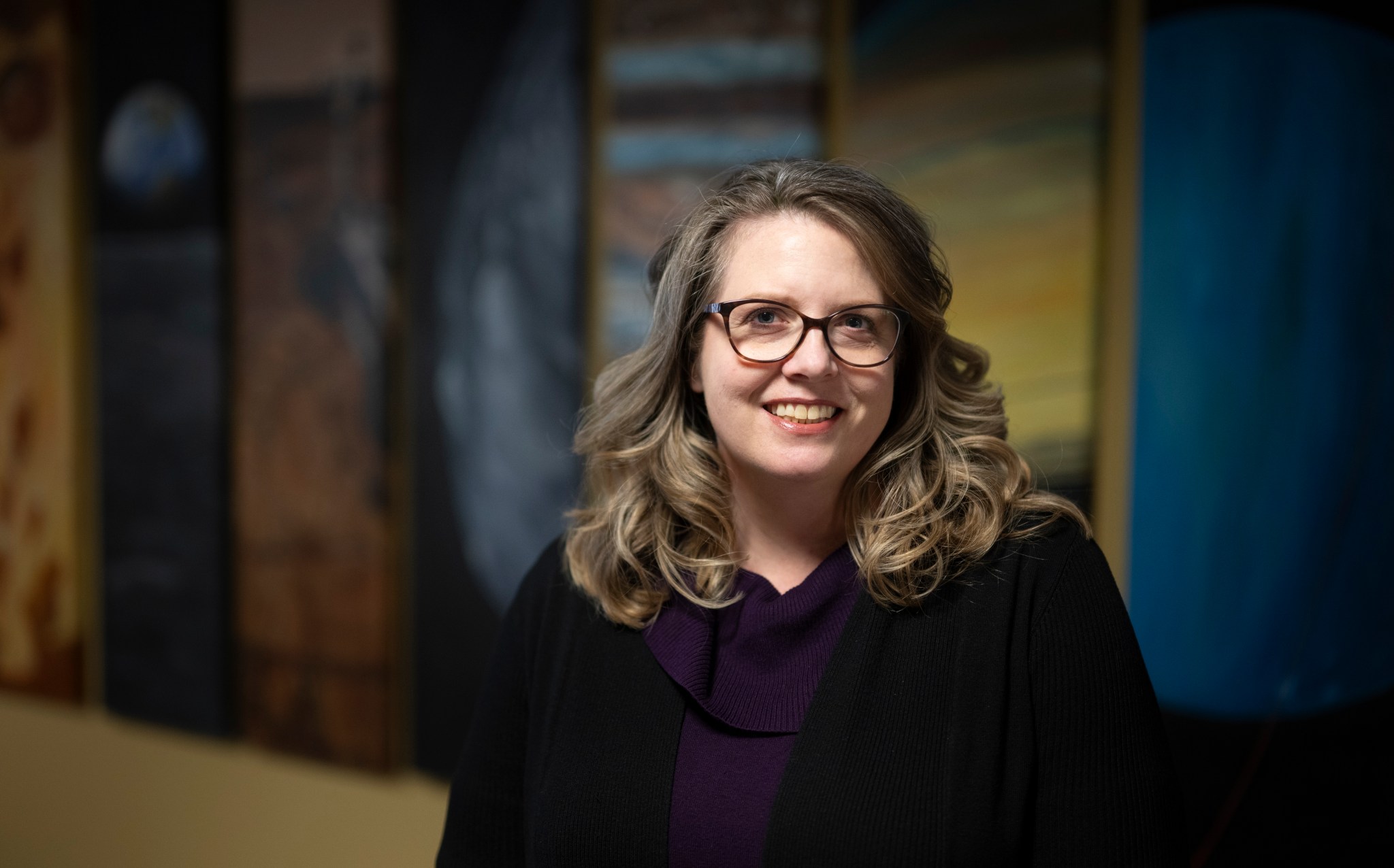 A woman wearing thick-rimmed glasses, a purple sweater, and a black cardigan smiles widely at the camera. Paintings of worlds across our Solar System are blurred in the background as light from above perfectly frames her face, surrounded by loose, dirty blonde curls. 