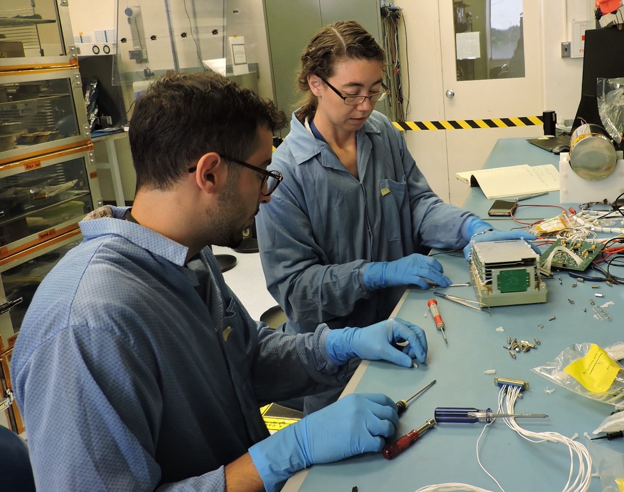 Two people seated at a workbench, wearing blue lab coats and gloves, and assembling an electronics board stack.