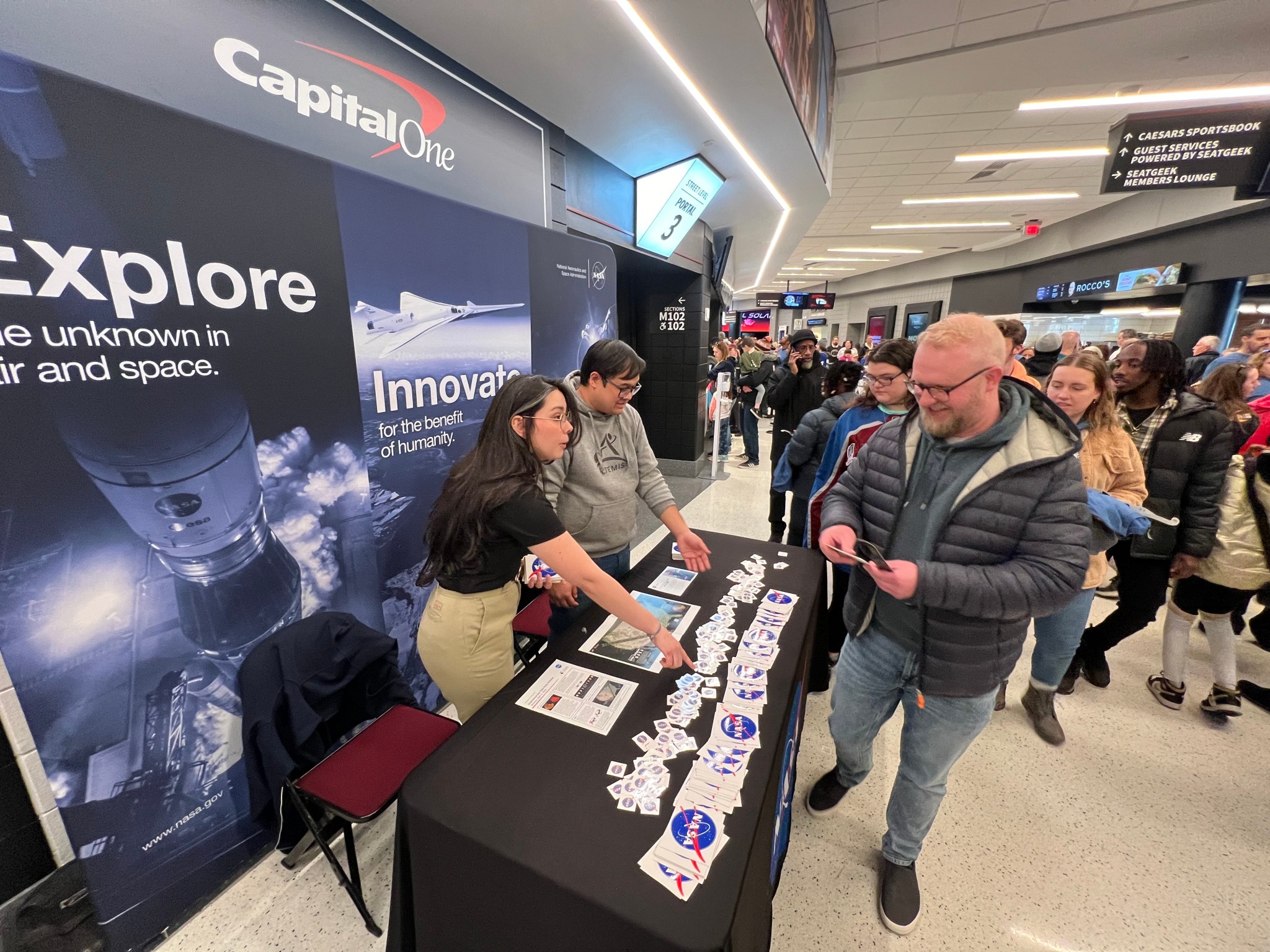 Two NASA representatives stand at the back of a table filled with NASA stickers, tattoos, and a map showing the path of the solar eclipse. People line up along the front of the table to talk with the representatives and take the NASA items.