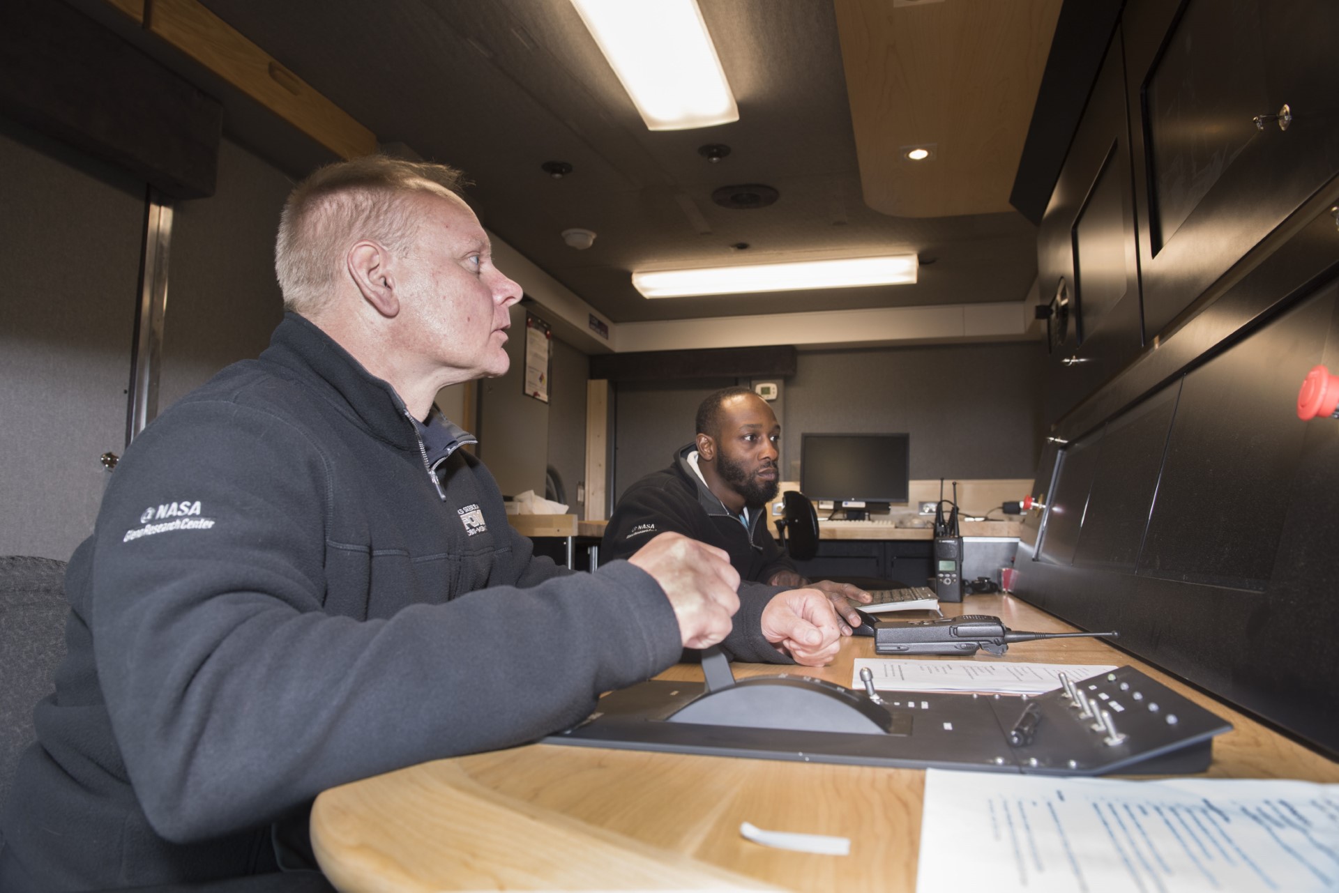 Two people sit at a console desk and supervise data from a jet engine test.