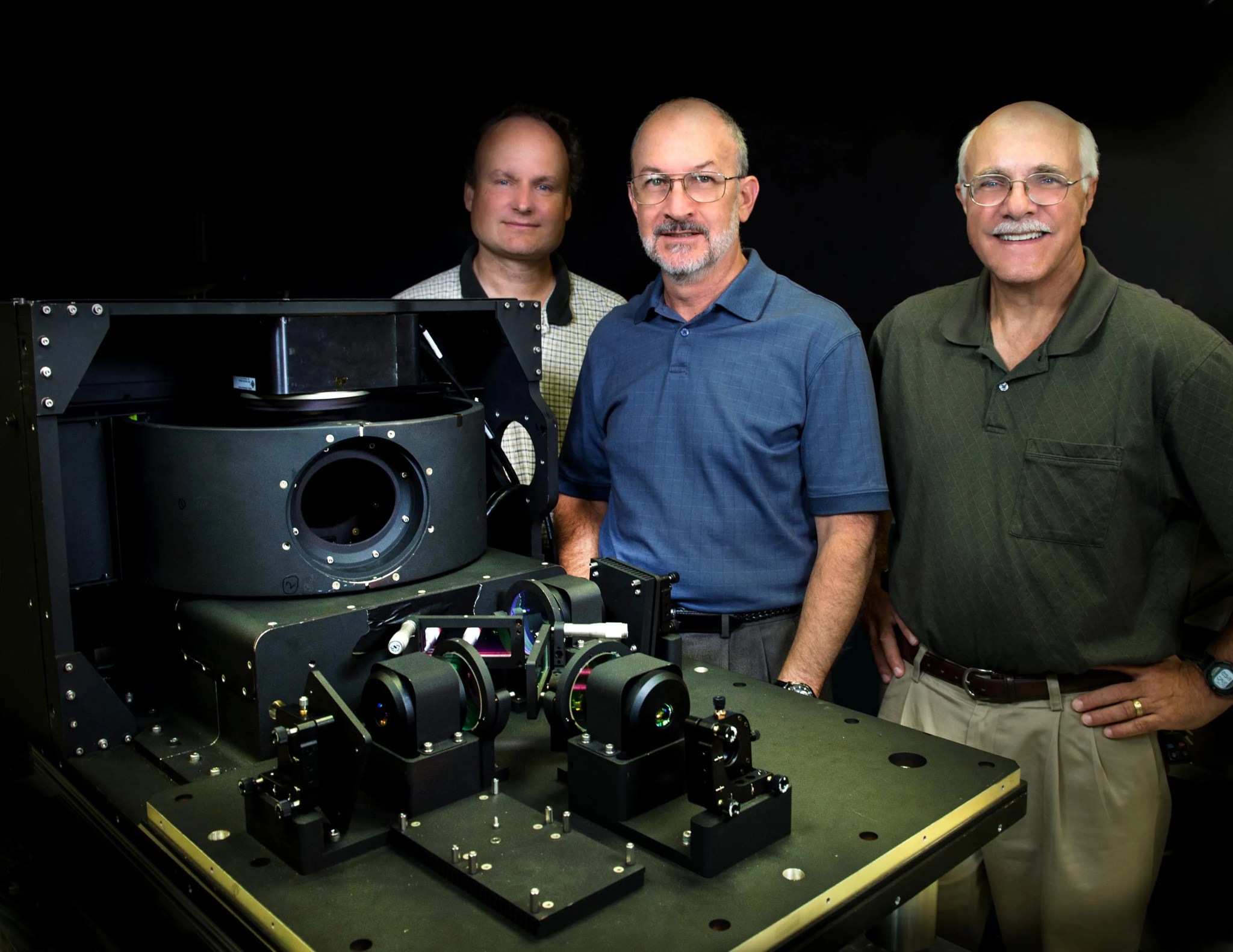 three men standing beside a small, black piece of space satellite hardware