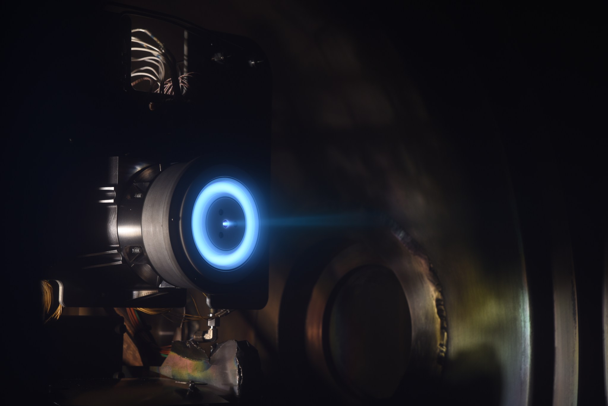 A cylindrical metal device with an azimuthal white channel mounted to a metal support structure inside a much larger metal chamber. A blue glow lights up the azimuthal chamber and a blue plume radiates light to the right of the thruster.