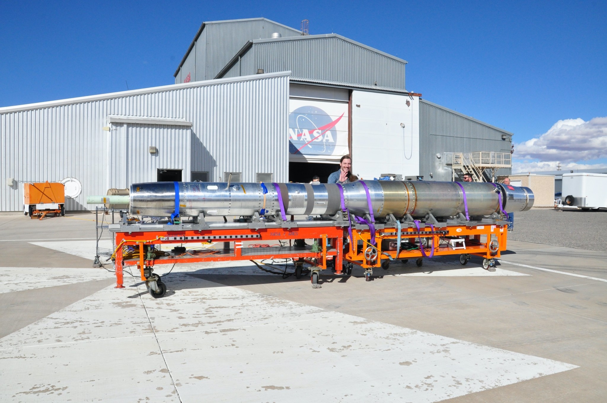 Austin Bumbalough, an electronics engineer at NASA’s Marshall Space Flight Center, waves from behind the Hi-C payload in front of the Vehicle Assembly Building in White Sands, New Mexico, in February 2024. The payload will be used in the Hi-C rocket experiment planned to take place sometime in April.