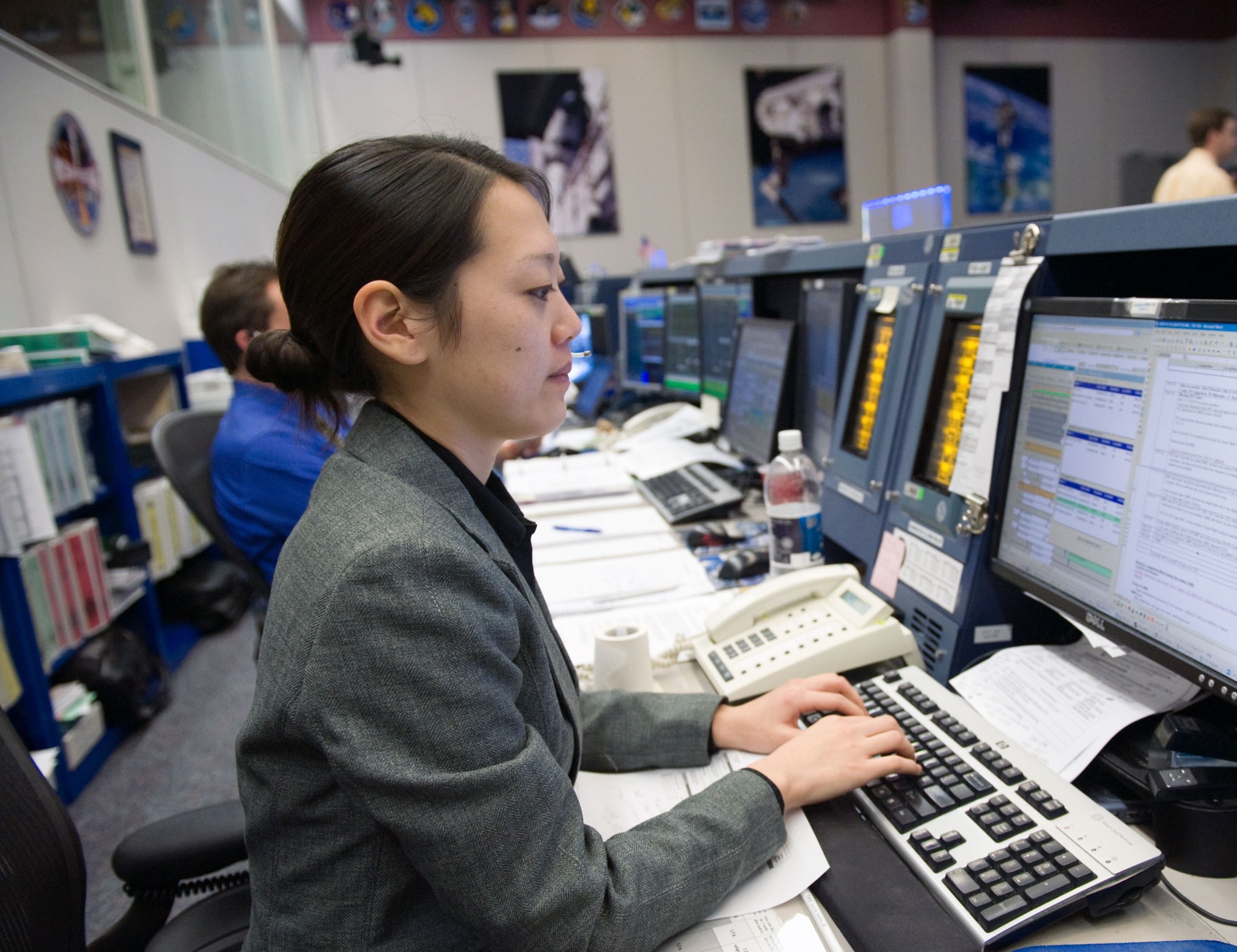 A woman wearing a grey suit with her hair tied back sits at a desk and works on her computer.