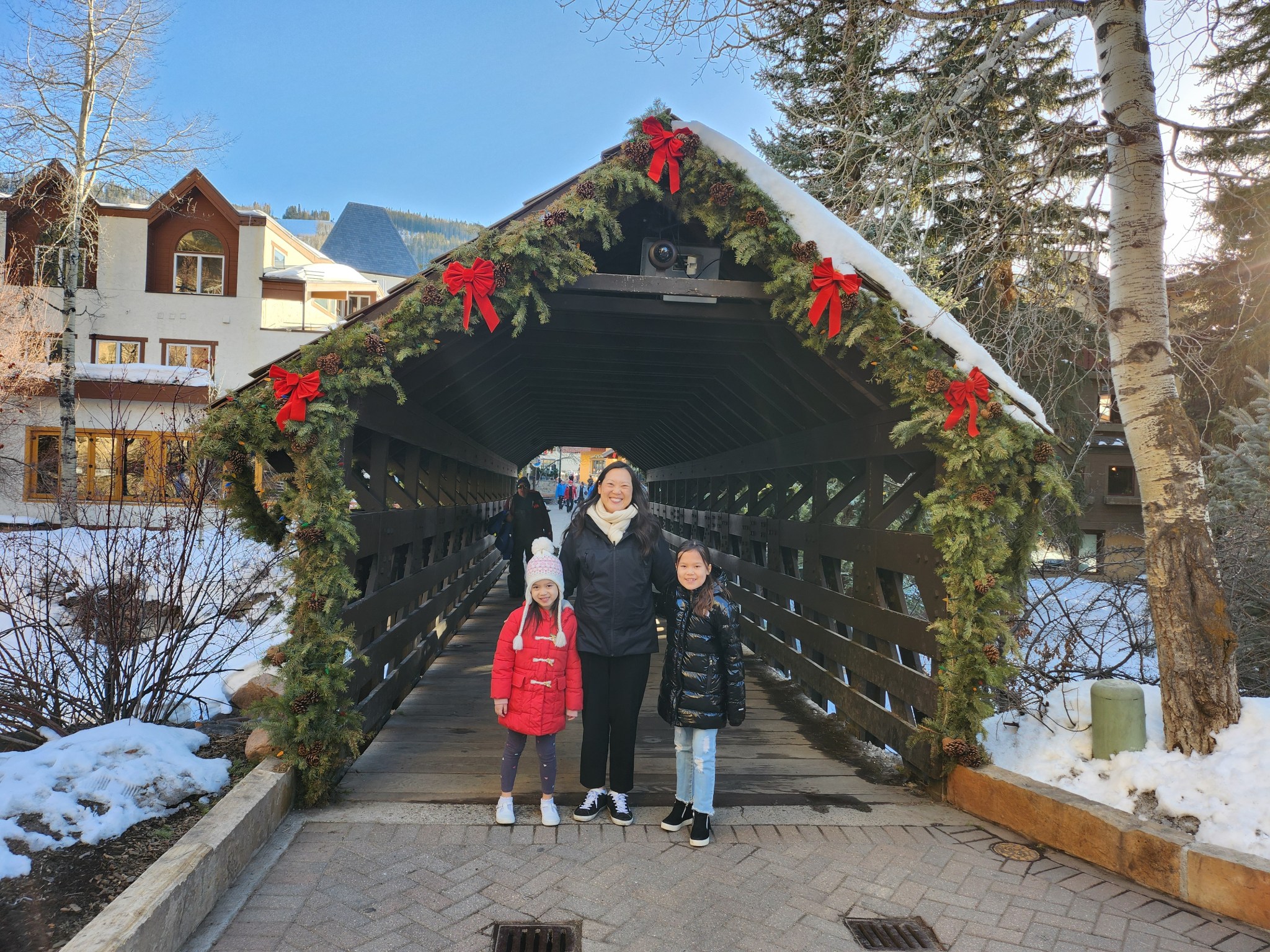 Woman and two young girls stand in winter clothes outside in front of a bridge.