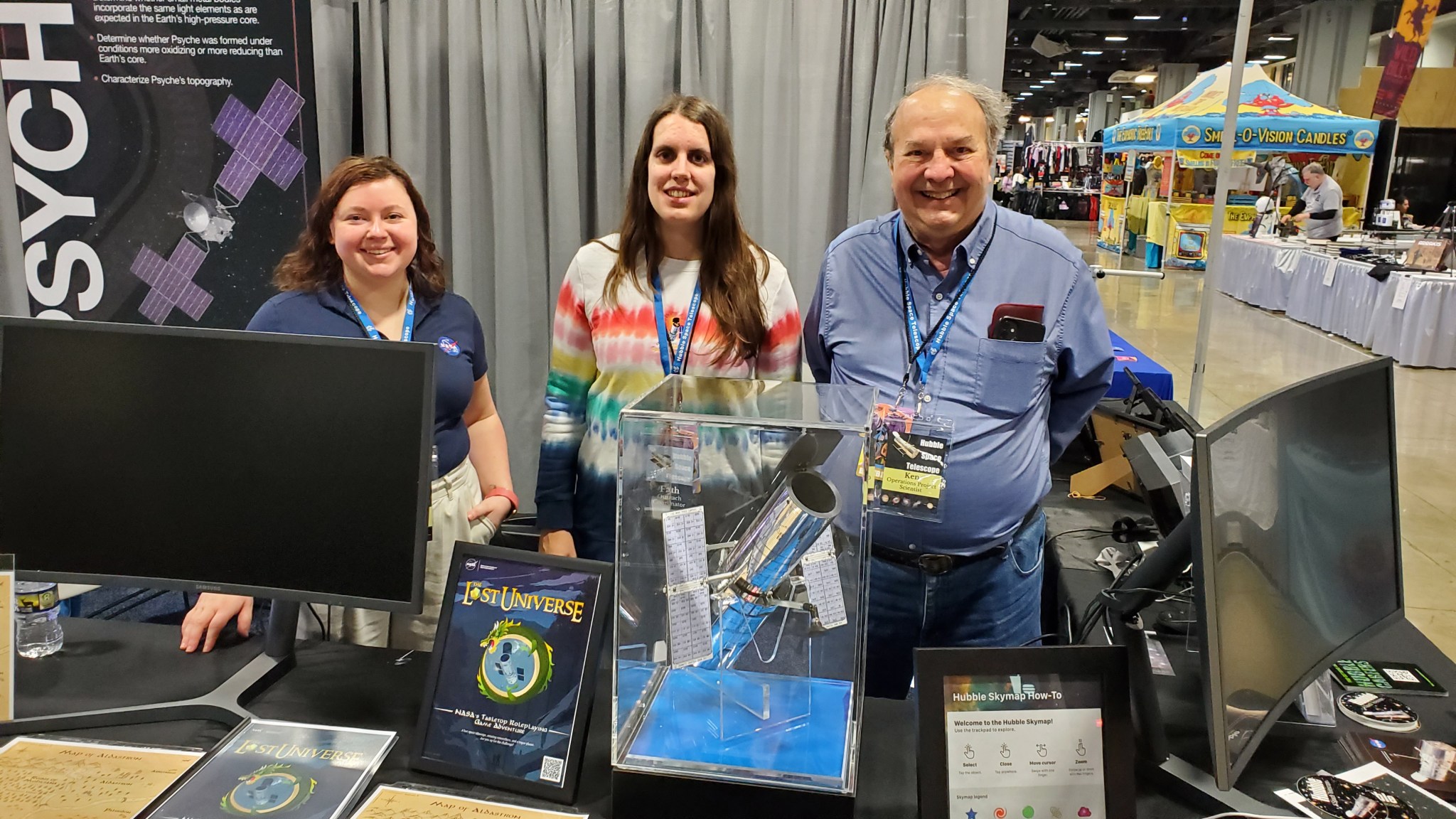 Three people stand at a booth at a conference with a Hubble model on the table and a sign for the "Lost Universe" table top role playing game. 