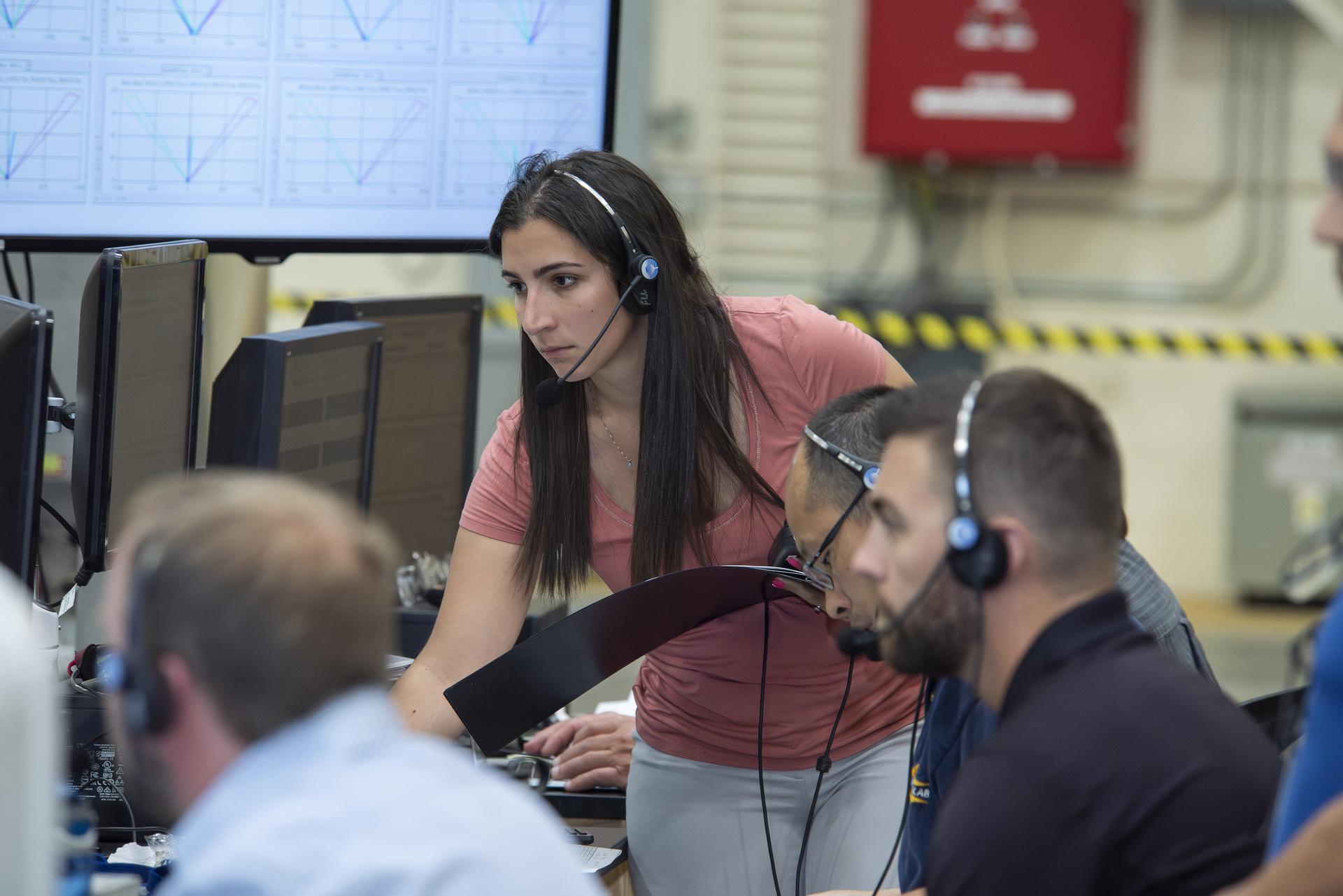 A woman hovers over a desk with multiple computer screens, wearing a microphone headset and holding a folder in her hands. Three men out of focus are seated in a row observing the multiple monitors.