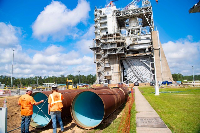 crew members are seen standing near new pipeline sections. the Fred Haise Test Stand is seen in the background