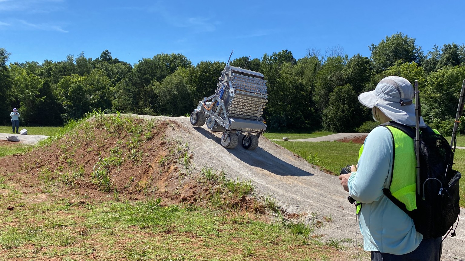 A man in a yellow vest, hat, and backpack uses a controller to drive his robot up a 30 degree incline outdoors.