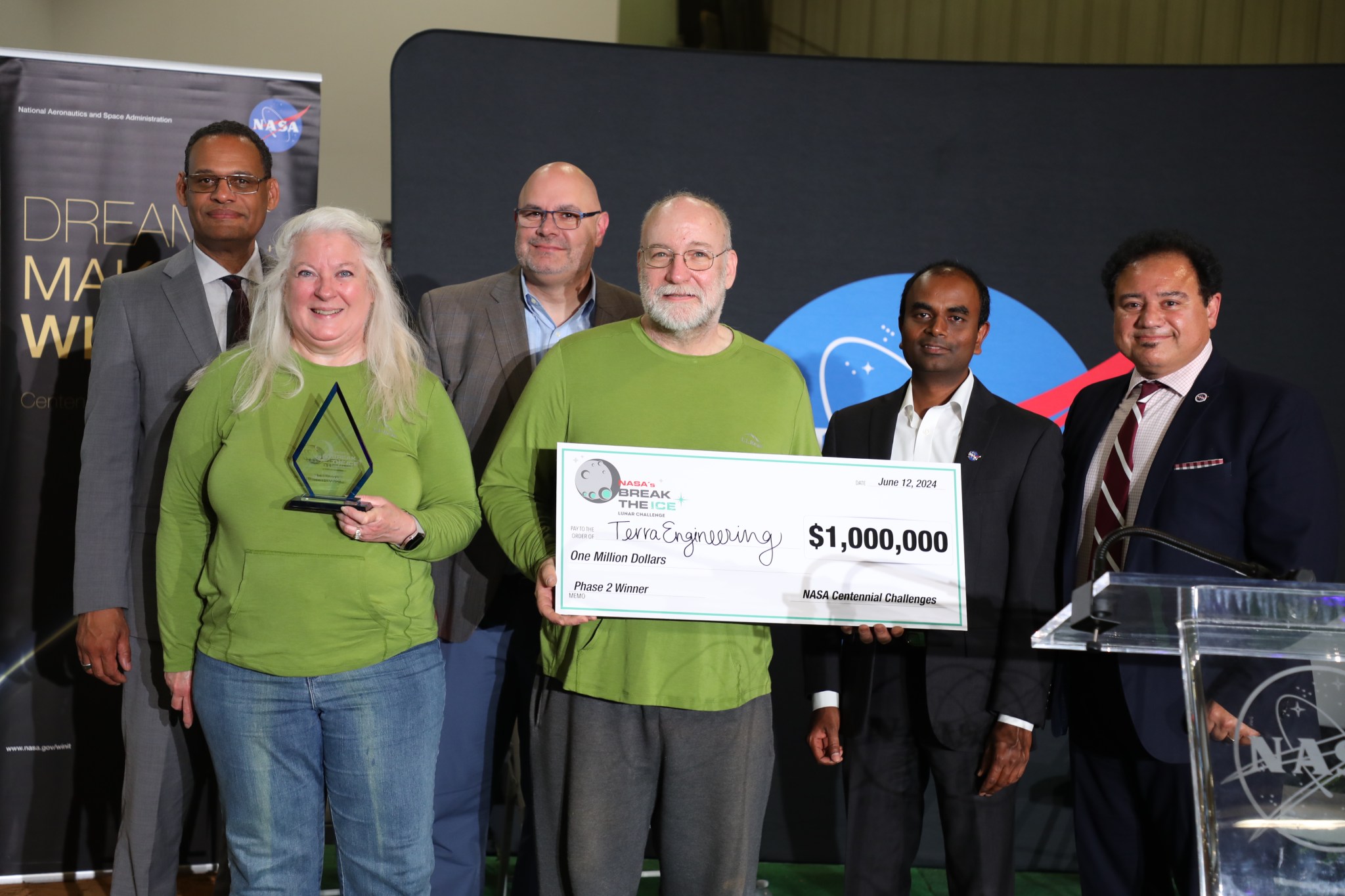 A older man and wife stand in green shirts holding a large check for a million dollars after winning NASA's Break the Ice Lunar Challenge