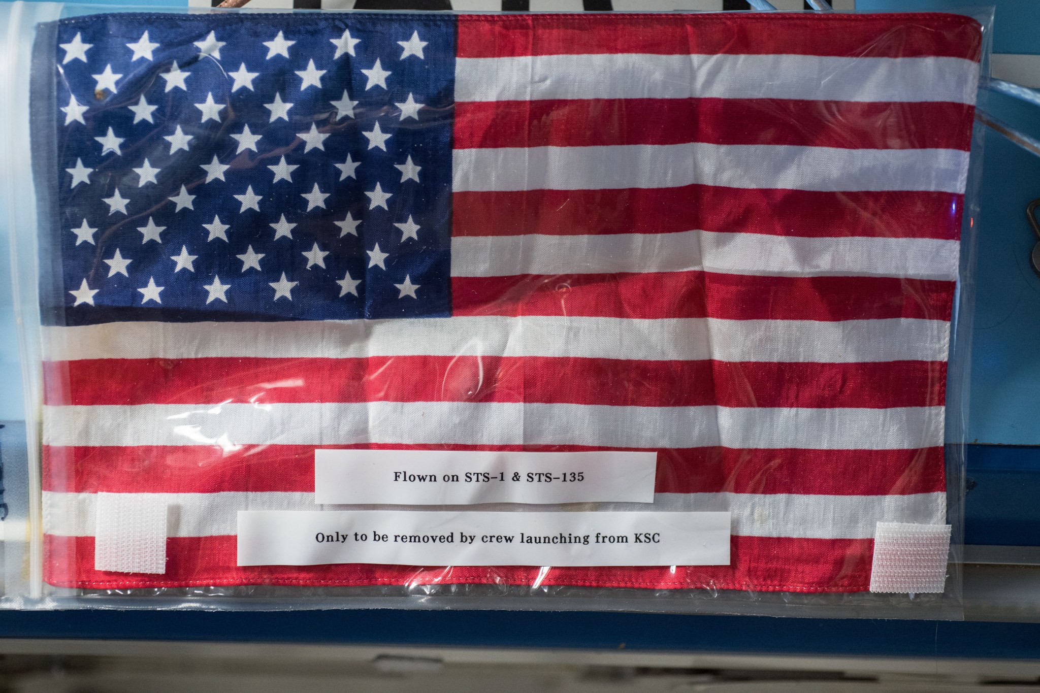 An American flag in a plastic bag with the words "Flown on STS-1 & STS-135, Only to be removed by crew launching from KSC" on the outside of the bag.