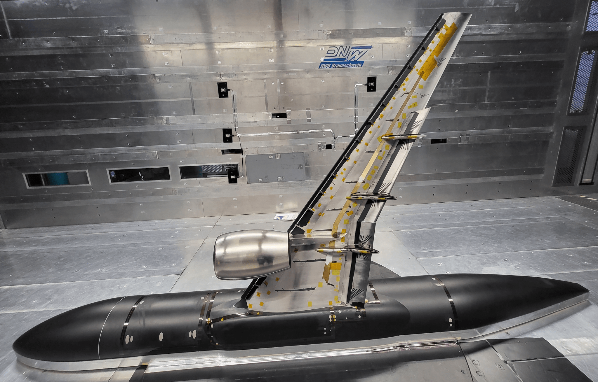 A silver-colored airplane research model sits in a wind tunnel facility. The model consists of a section of a scaled-down fuselage on the floor, with a single wing mounted vertically on it. The metallic wall of the wind tunnel is visible in the background.