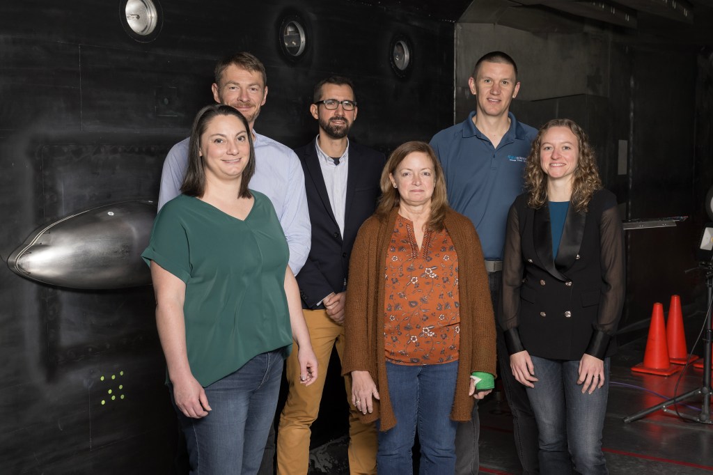 Six researchers stand in front of a sliver-colored airplane research model installed in the wind tunnel.