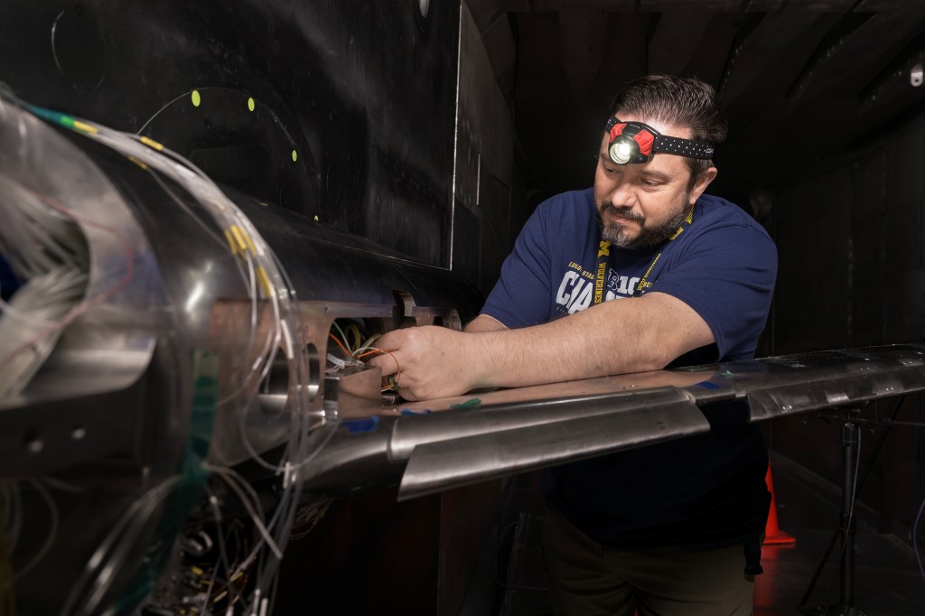 A technician wearing a head lamp is working on the wiring that is inside a sliver-colored airplane research model.