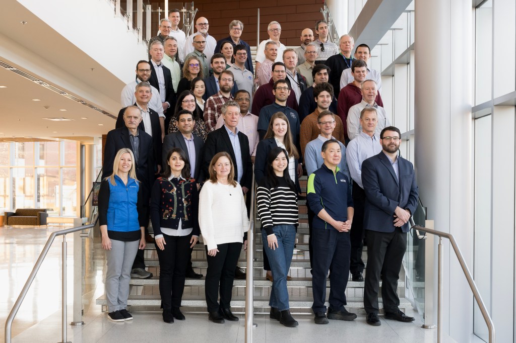 A group of 42 researchers stand on a staircase inside a building to pose for a group photo.