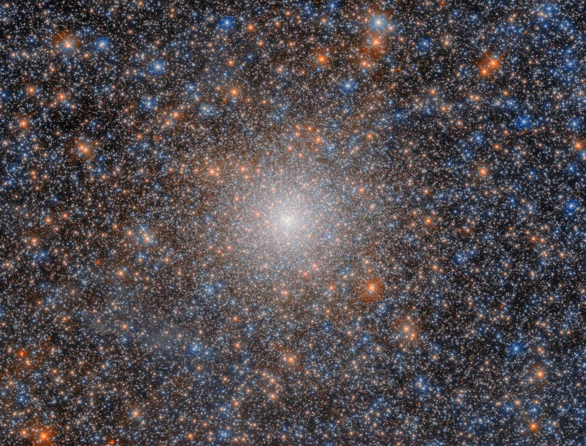 A globular cluster that looks like a very dense, ball-shaped collection of many shining stars in colors of white, yellow-orange, and blue. Some stars appear a bit larger and brighter than others, with the brightest having faint cross-shaped diffraction spikes. The cluster’s stars are scattered mostly uniformly, with their density increasing toward the cluster’s core where they merge into a strong, bright-white glow.