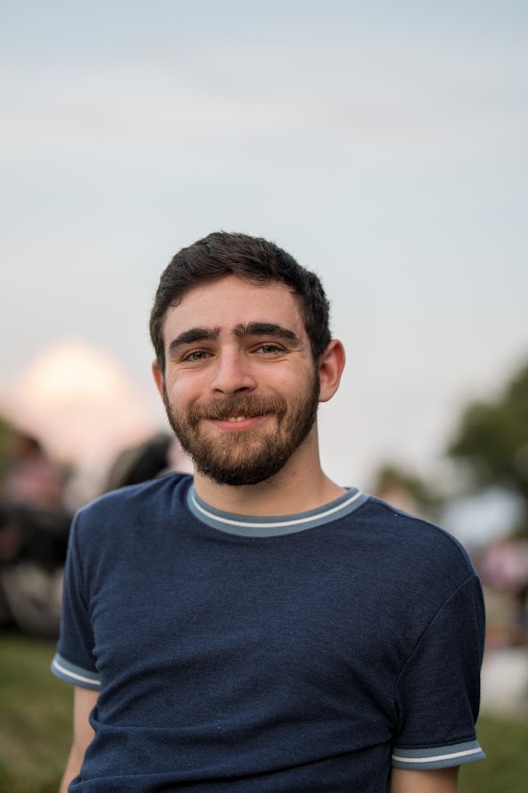 Jake Cupani stands outside, smiling, wearing a navy blue t shirt. The background is out of focus and includes a blue sky, grass, and trees. 