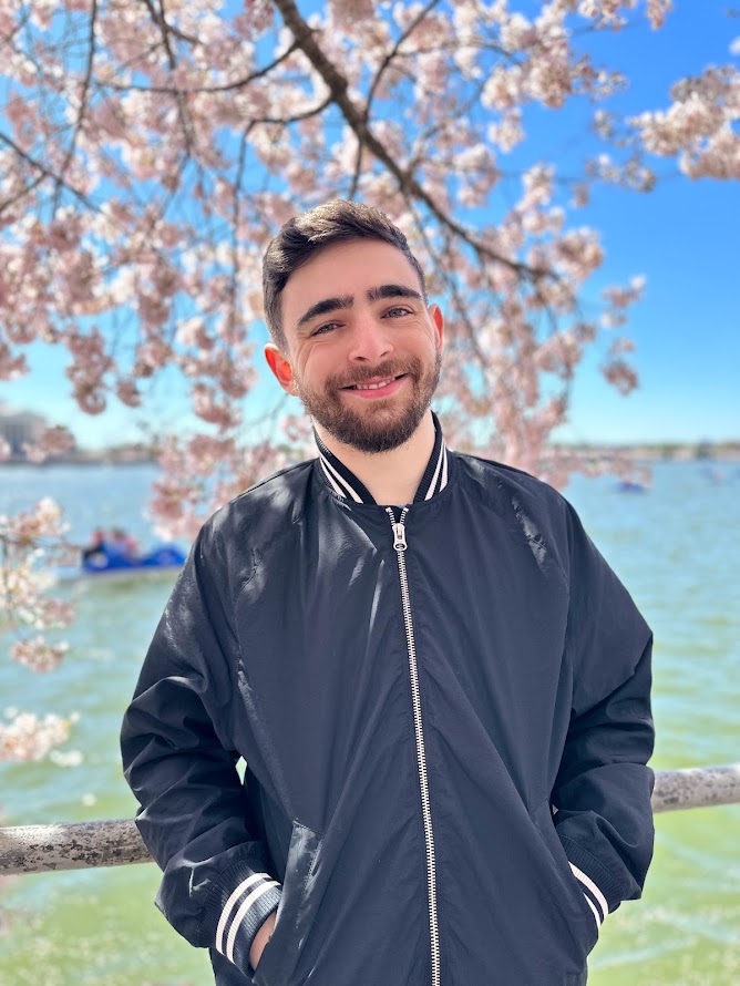 Jake Cupani smiles in front of a body of water with a cherry blossom tree directly behind him. He has short brown hair and is wearing a navy blue jacket. His hands are in his jacket pocket. 