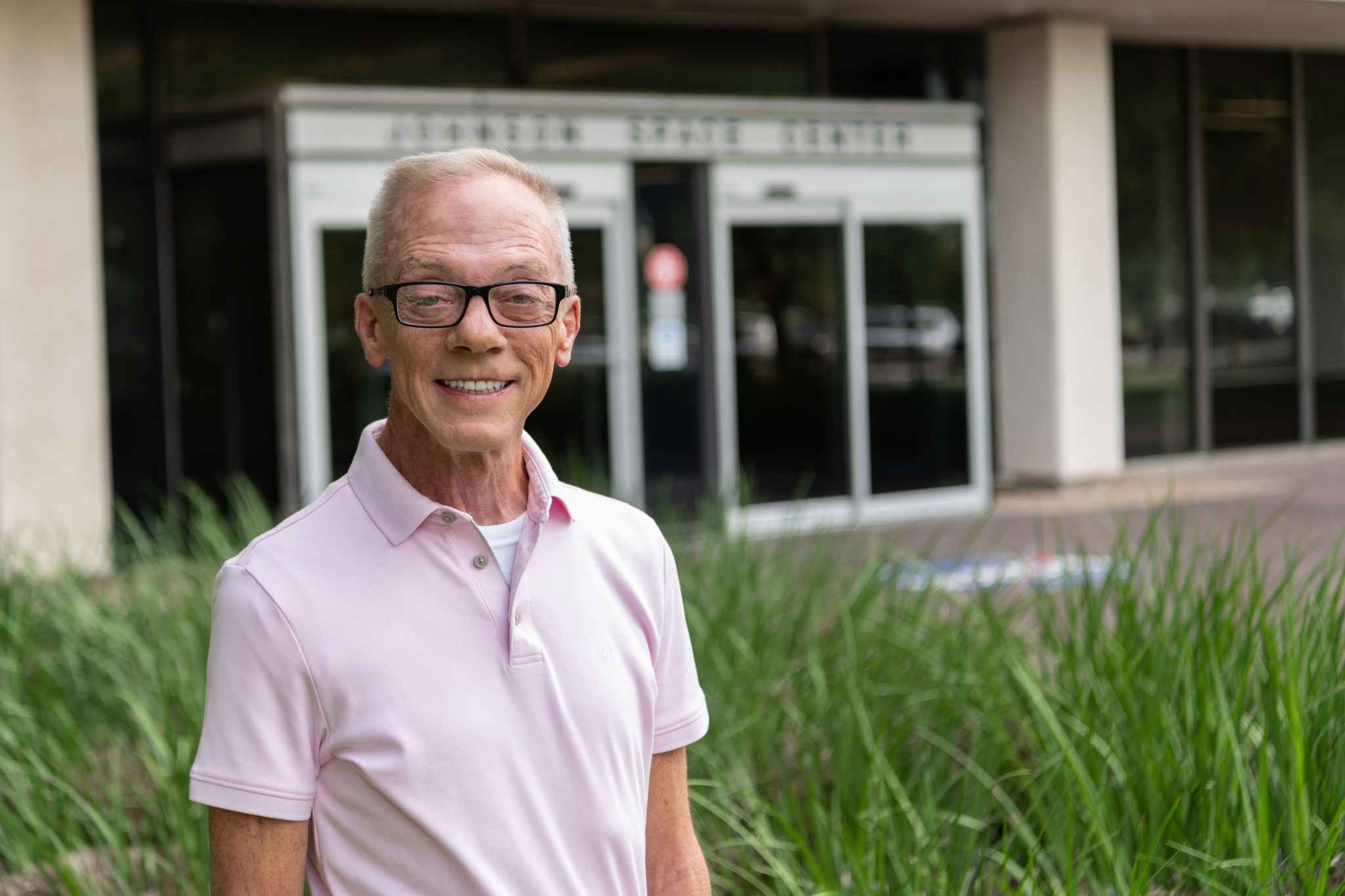 A thin white man with thick-rimmed glasses and wearing a light pink polo shirt stands in front of an office building with Johnson Space Center written over the door.