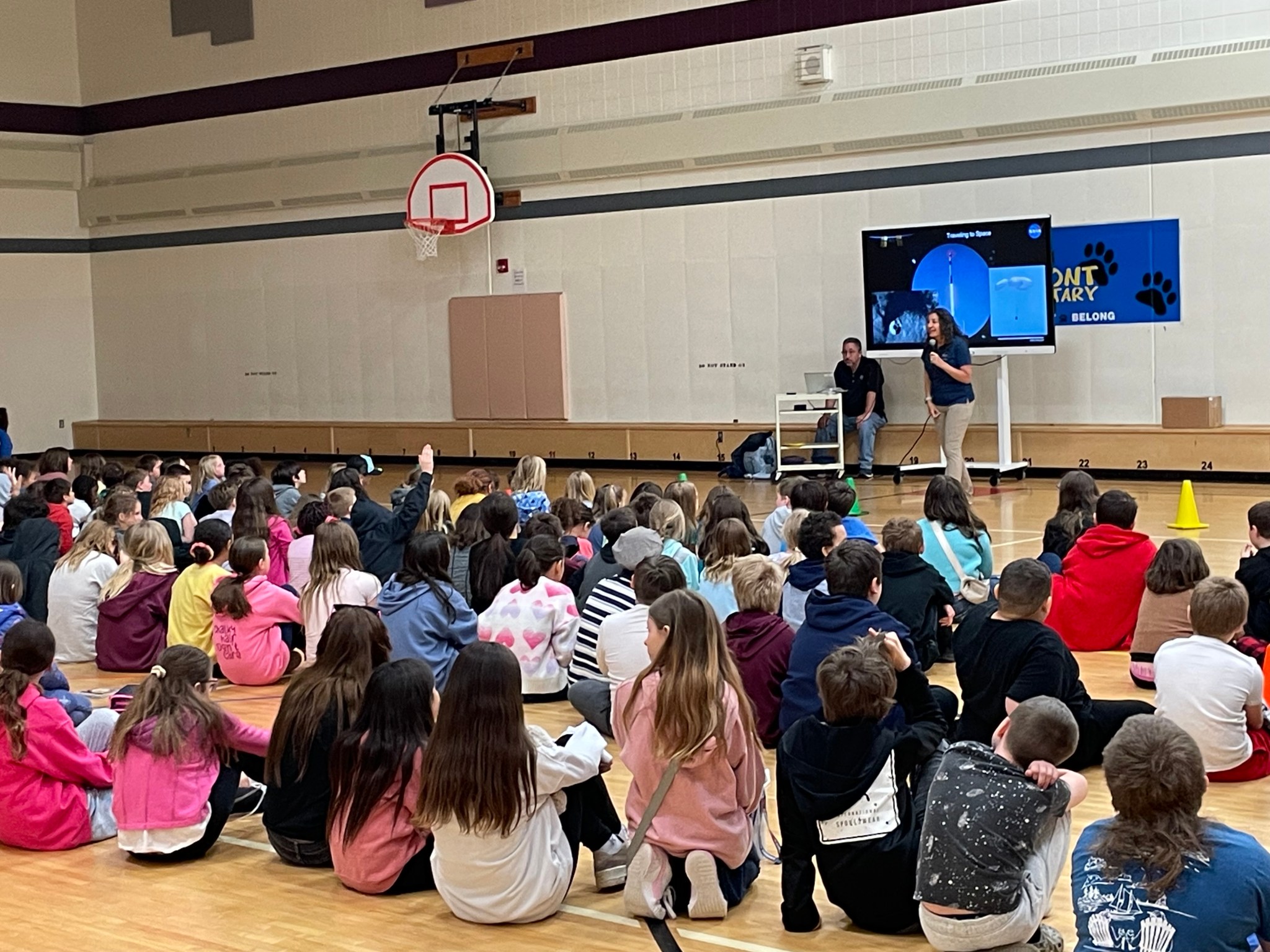 Astronaut stands in front of a gym full of grade-school students seated on the floor and shares her experiences on the International Space Station. A large screen in the background shows highlights from space.