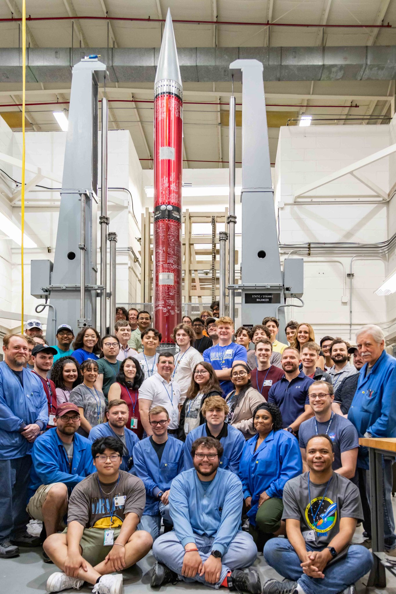 A group of smiling students sit in front of a red rocket that is standing vertically in a balance test behind them.