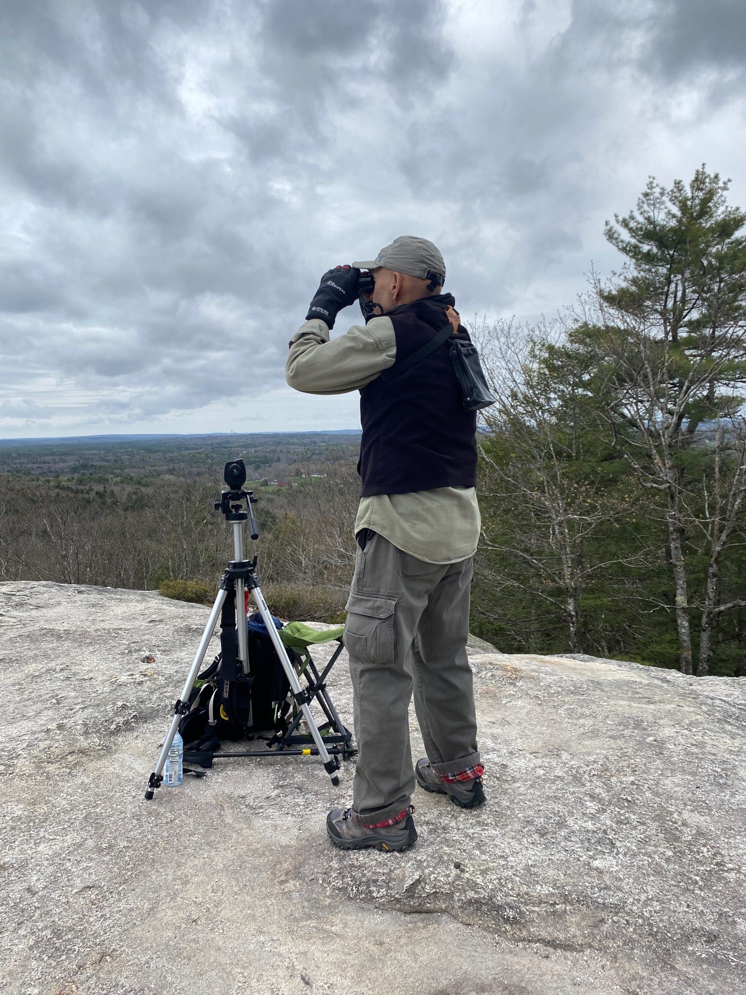 A man standing on a large rock overlooking a valley. The man is visible from behind looking through binoculars. A tripod is in front of him. 