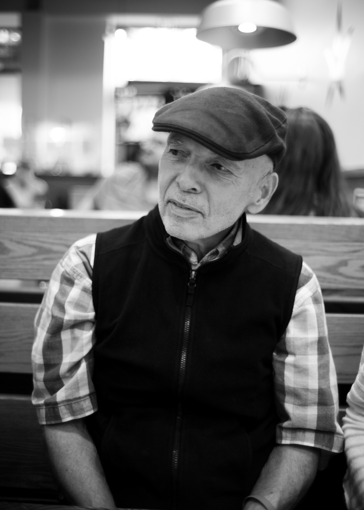 Black and white image of a man sitting on a wooden bench wearing a hat, vest, and plaid short sleeve shirt. 