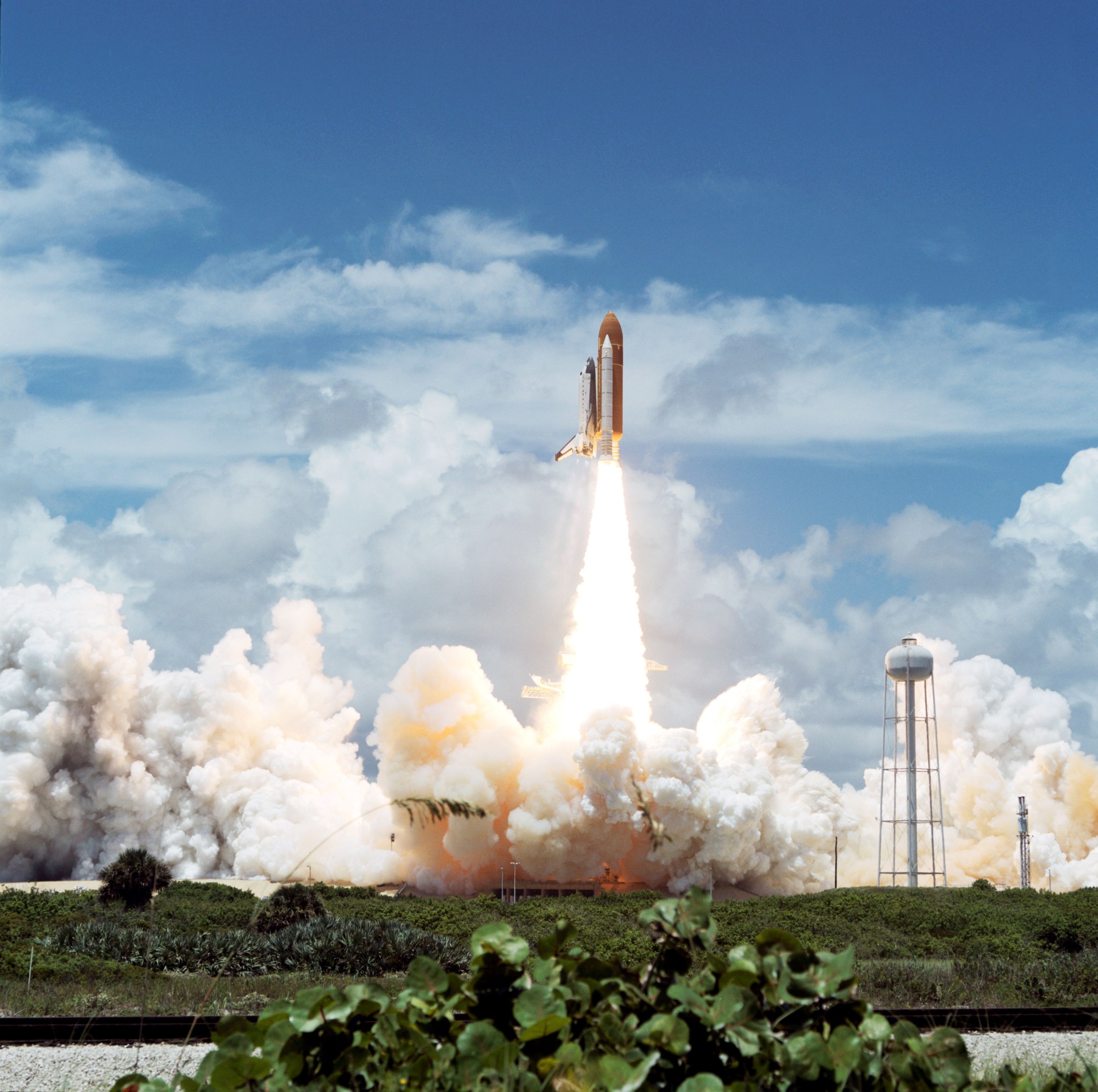 Space shuttle Columbia lifts off from the launchpad, heading upward through the partly cloudy blue sky. A conical spray of fire comes out from the bottom of the spacecraft. It is the brightest thing in the image. Below that, clouds of vapor spread out horizontally across the pad. In the foreground are some green plants.