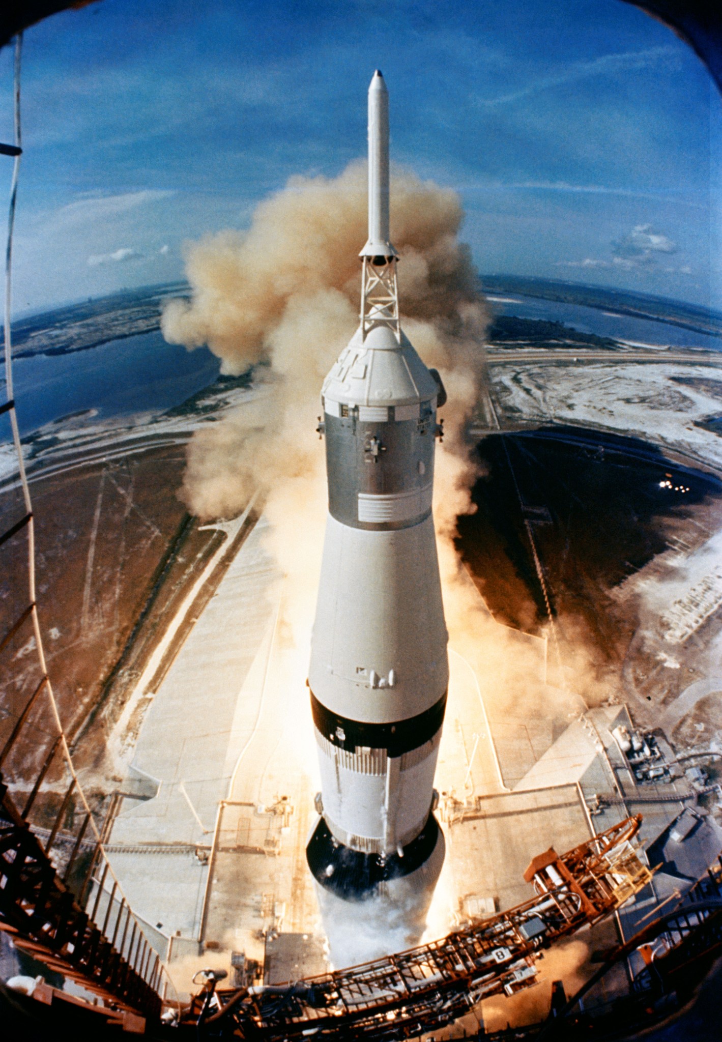 The Apollo 11 spacecraft lifts off from a launchpad. The view looks downward at an angle, so the entirety of the spacecraft is visible. Vapor spreads out behind the rocket. Further in the distance is blue water and sky.