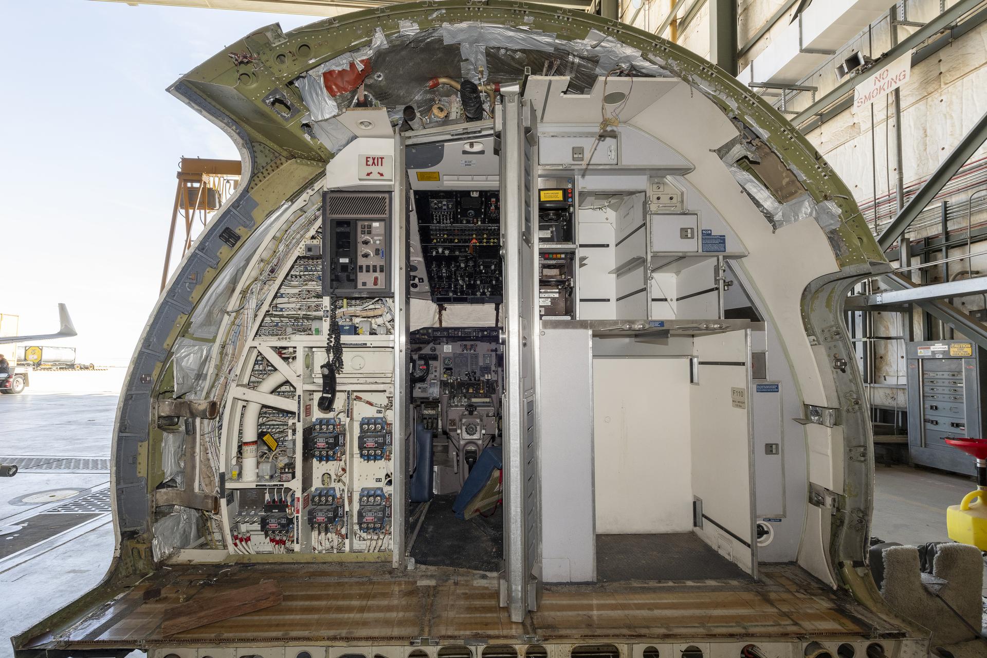 Part of the cockpit of an airplane is seen in the center of the photo, there are wires and panels visible throughout, the background of the photo is full of light from an open hangar door.