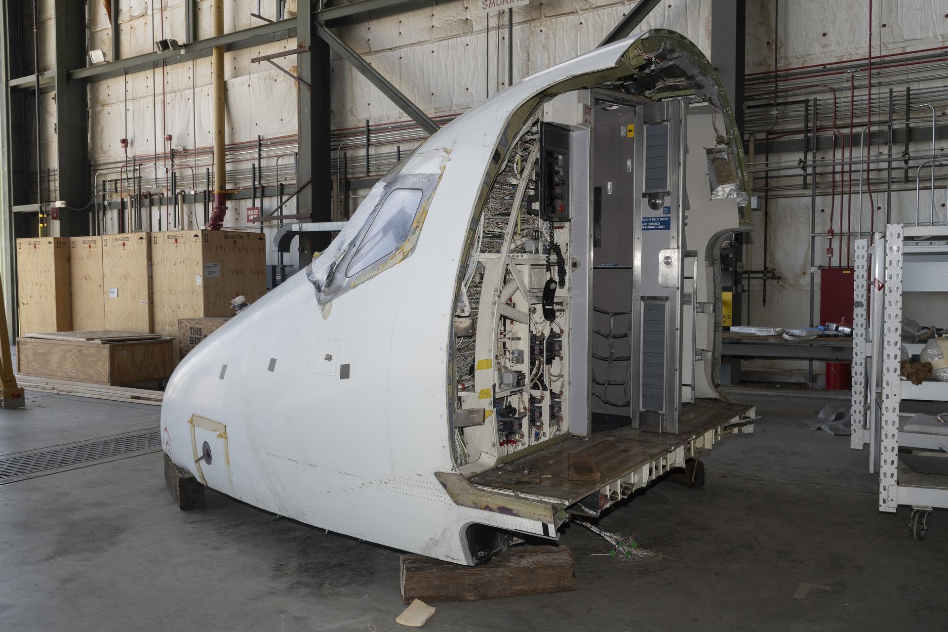 Photo of the front part of an airplane in the center, with a concrete ground under, the cockpit is sitting on wood blocks, there are wires and panels through the part of the aircraft that is visible as well as behind the cockpit. 