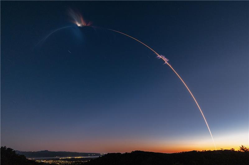 A photo taken with extended exposure shows the streak of light made by a rocket in a twilight sky. City lights are below on the left and the horizon has an orange glow where the sun has set in the lower right.