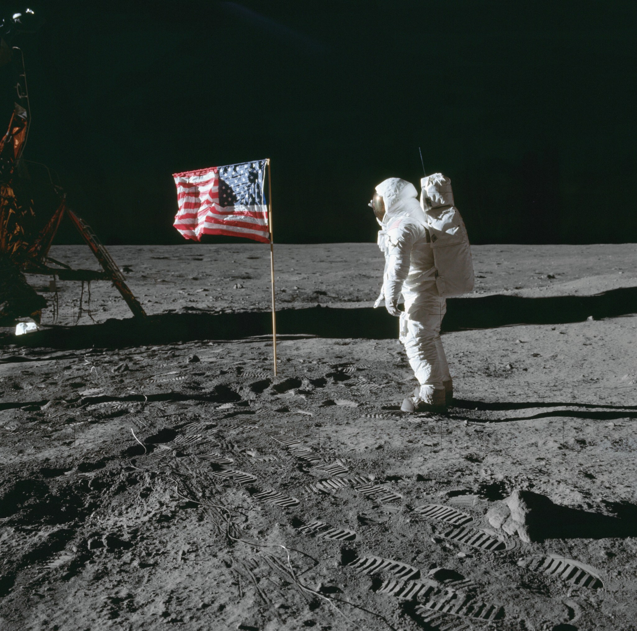 Astronaut Buzz Aldrin looks at a United States flag as he stands to the right of it. He is wearing a white spacesuit. The lunar module is on the left side of the flag. Bootprints in the gray lunar soil trail from the right in the foreground to the gold lunar module in the back. The darkness of space provides a contrasting backdrop to the image. The faint crosses on the image are markings that are used to calibrate distance and heights in photos taken on the lunar surface.