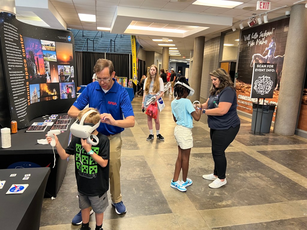 Young convention goers are assisted with the use of virtual reality devices by NASA employees