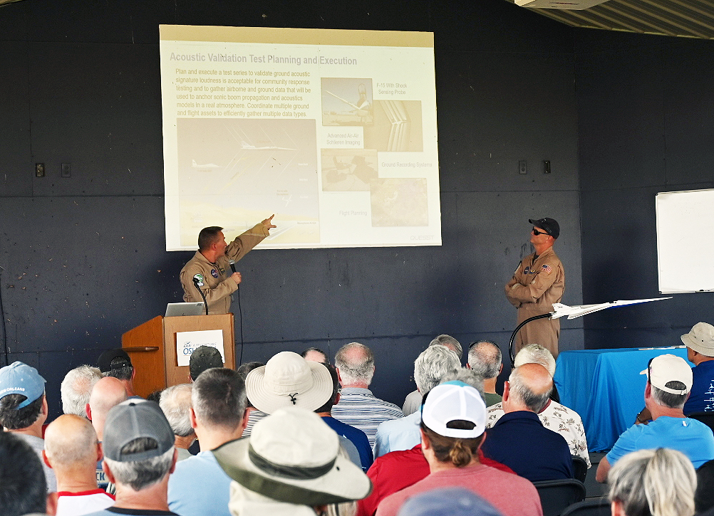 Two NASA test pilots stand behind a podium and an aircraft model while presenting a talk to large group of people in the foreground.