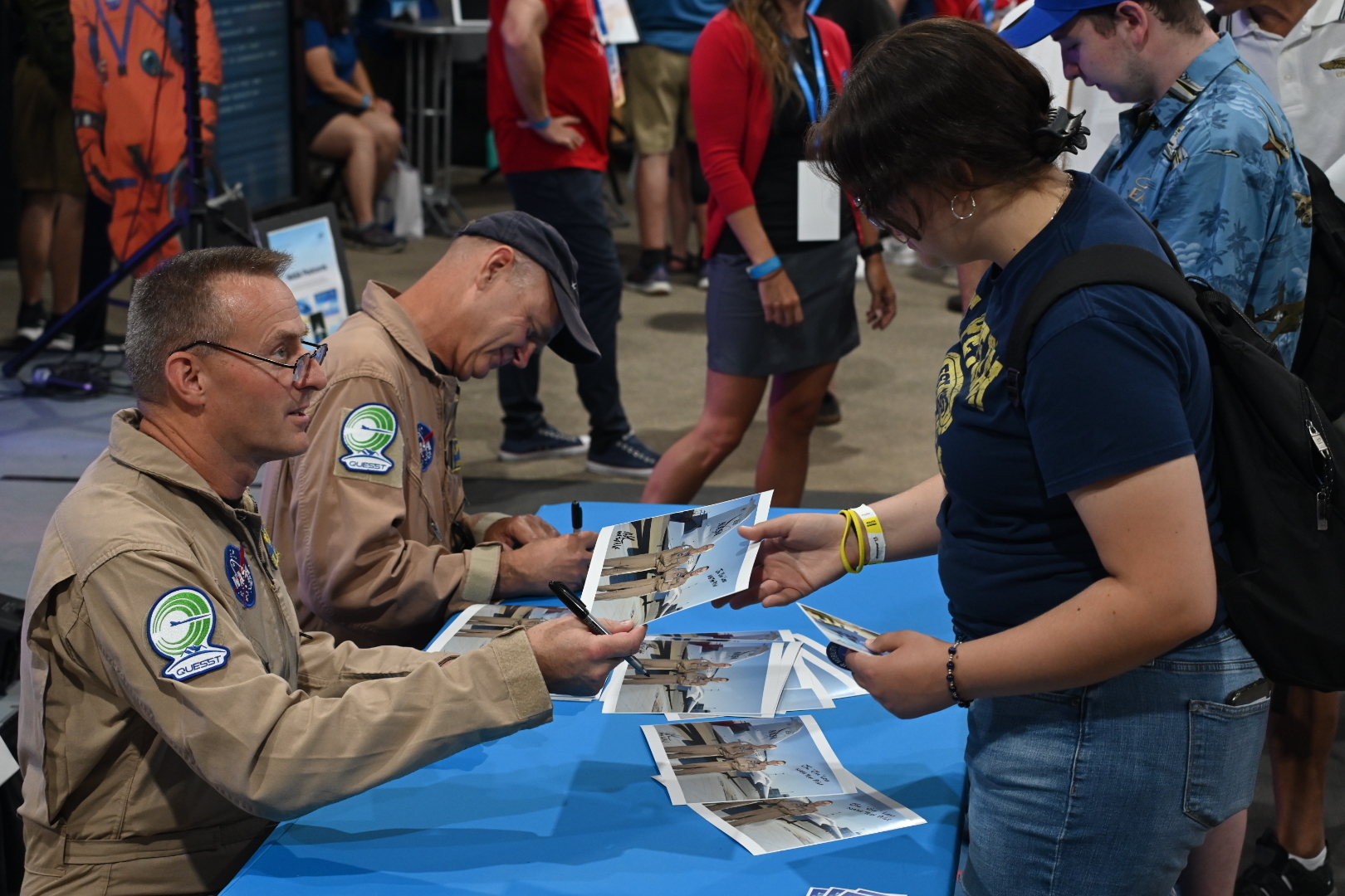 Two NASA test pilots sit at a table signing autographs for people attending Oshkosh 2024.