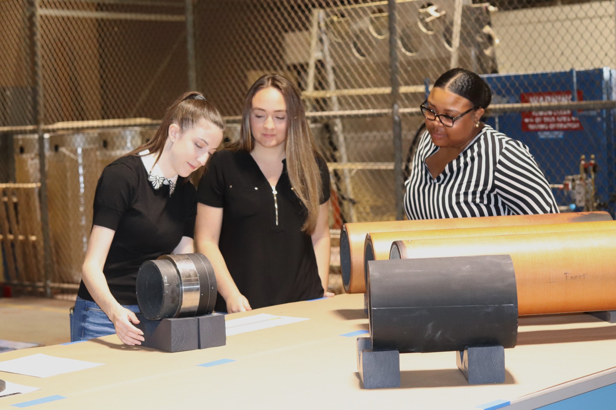 Three female NASA engineers conduct post-test analysis of disassembled, cylindrical components of a new, hybrid rocket motor testbed at NASA’s Marshall Space Flight Center.