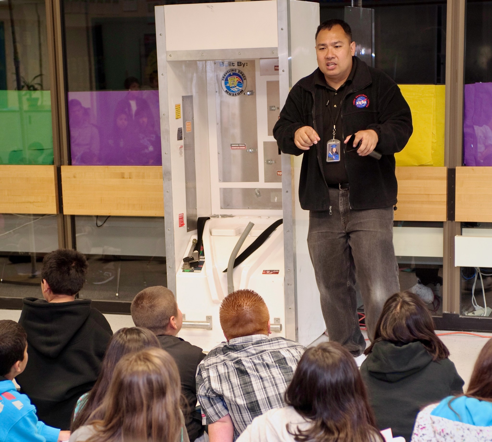 A group of students, seated on the ground, face the front of the room and listen while Jonas Dino talks to them.