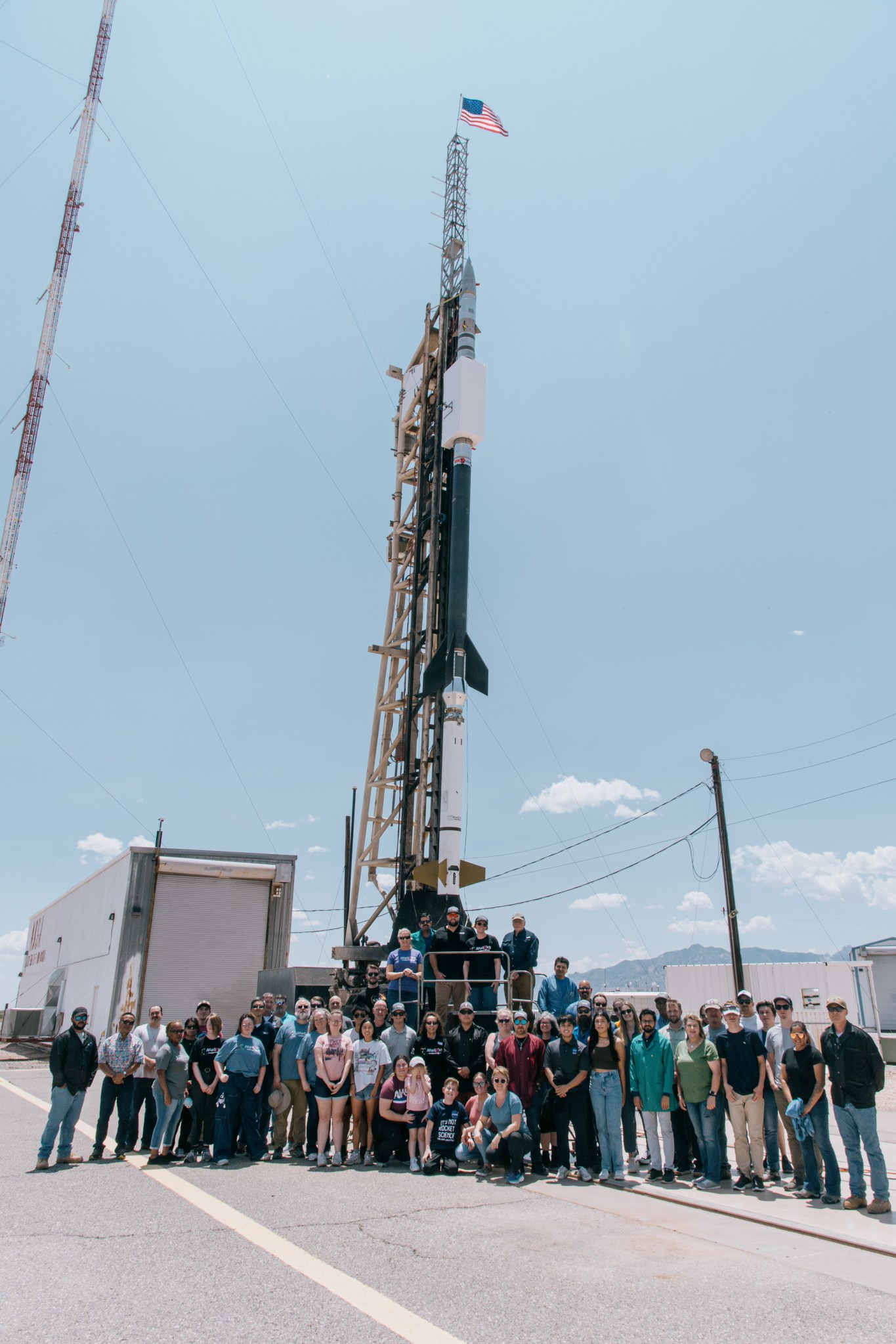 A group of around 30 individuals stand in front of a sounding rocket on a launch pad at a facility in White Sands, New Mexico.