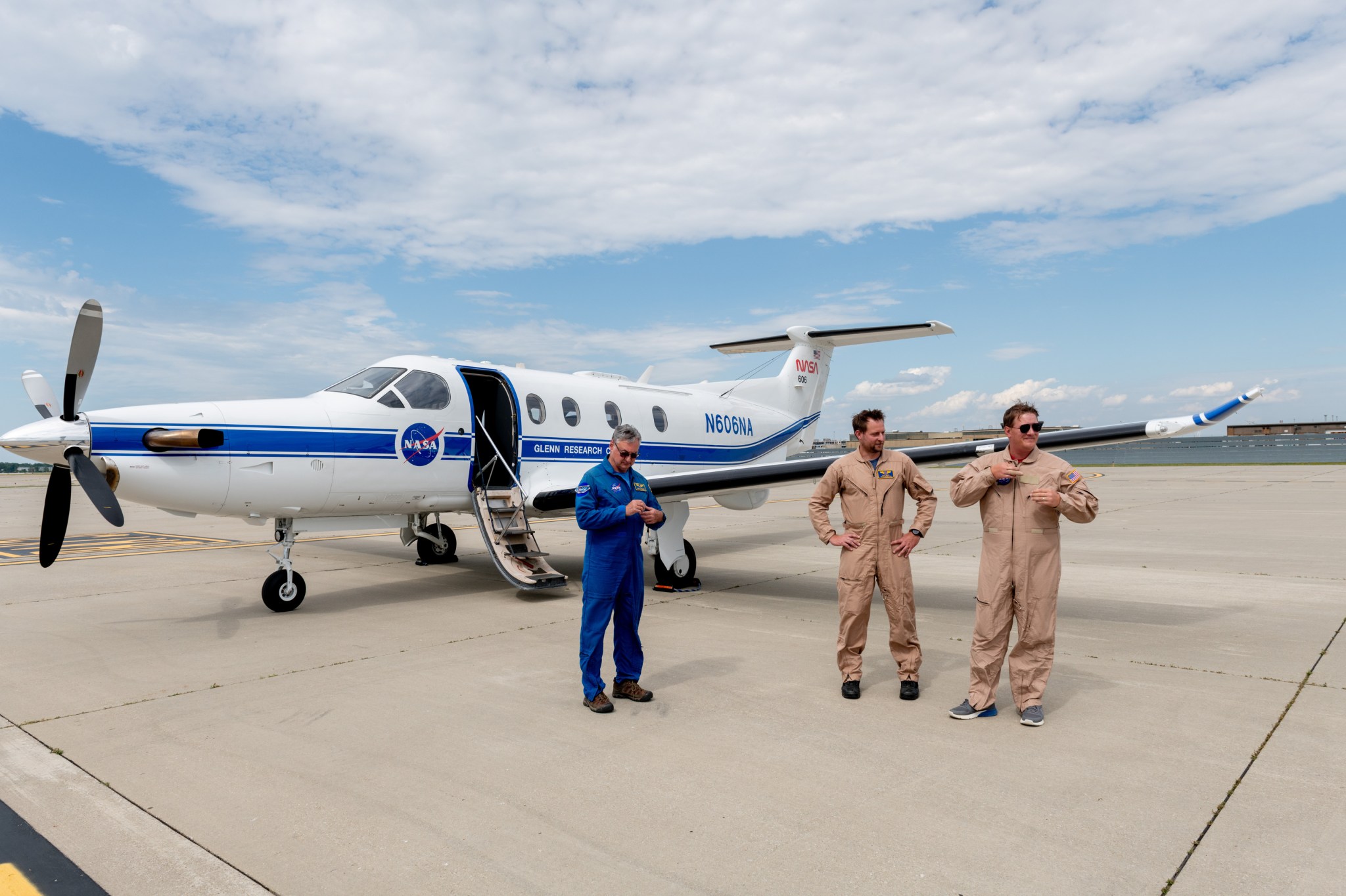 In this image we see the PC-12 aircraft sitting on a runway in front of a sunny blue sky filled with white clouds. The aircraft is shiny and white, with a blue stripe running across the side of the plane. The NASA meatball logo and the words “Glenn Research Center” can be seen on the side of the plane. Three men are standing in front of the aircraft, one wearing a blue NASA pilot’s uniform, while the other two wear tan flight uniforms.