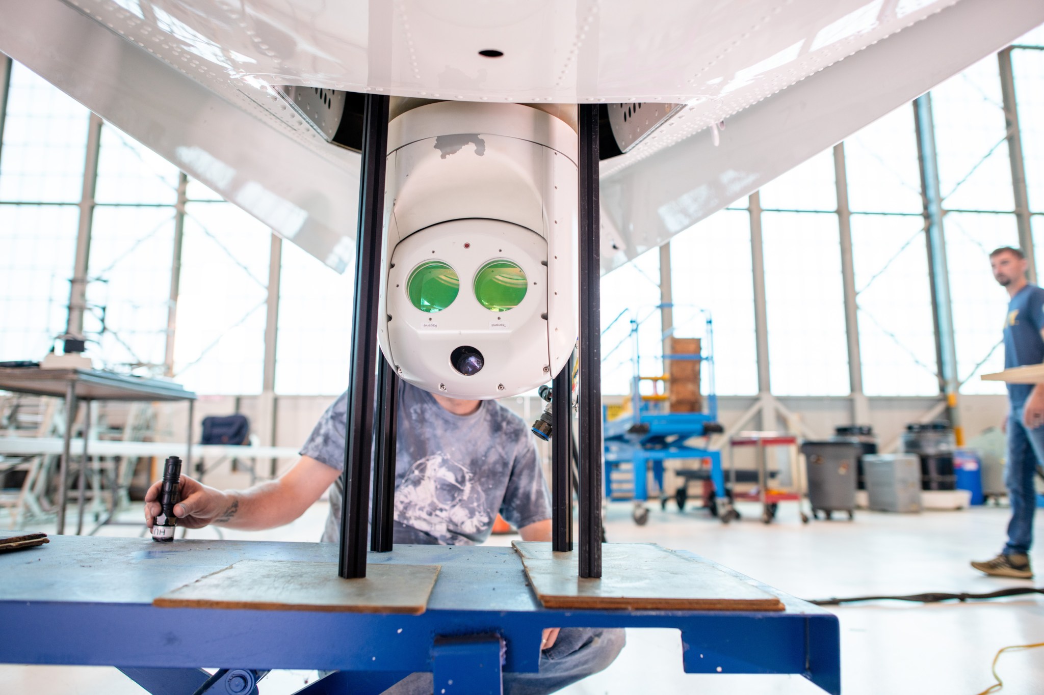 In this photograph of the white underbelly of the PC-12 aircraft, a white round optical system with two green mirrors is shown protruding out of a door on the bottom of the aircraft. Two men can be seen out of focus working in the flight hangar in the background of the image.