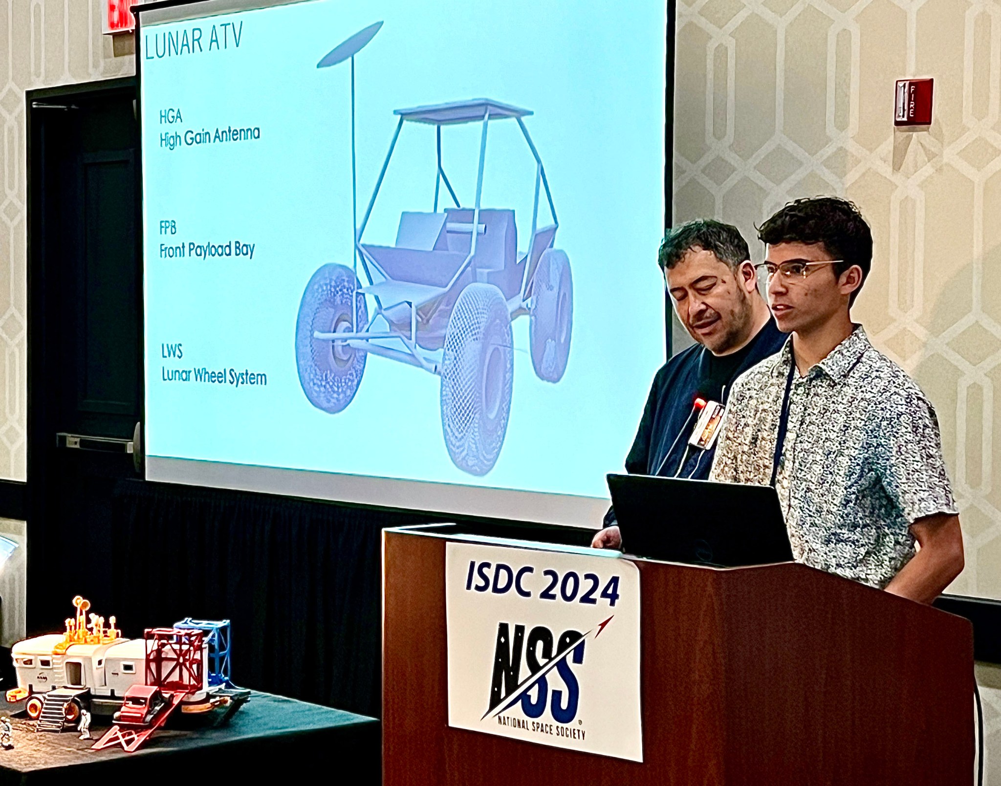 Finn Braun stands behind a lectern and to the left of Dr. Pascal Lee. Both are in front of a slide showing their design for a lunar ATV. A model of a lunar module is in front of them, on a table.