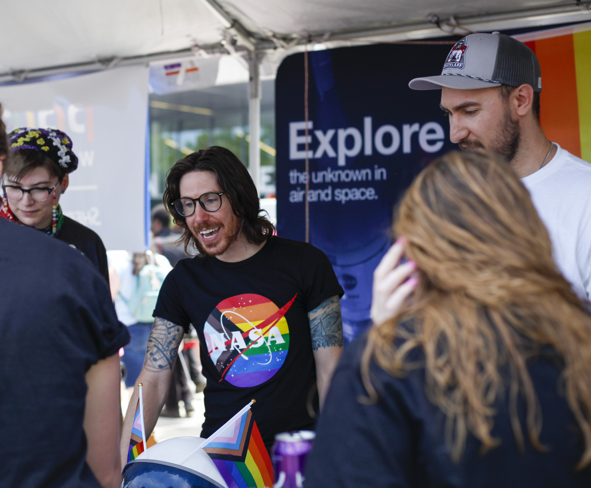 Three NASA Glenn Research Center employees talk with visitors at a table featuring rainbow flags and a NASA space helmet. A colorful banner reading, “Explore the unknown in air and space” hangs in the background.